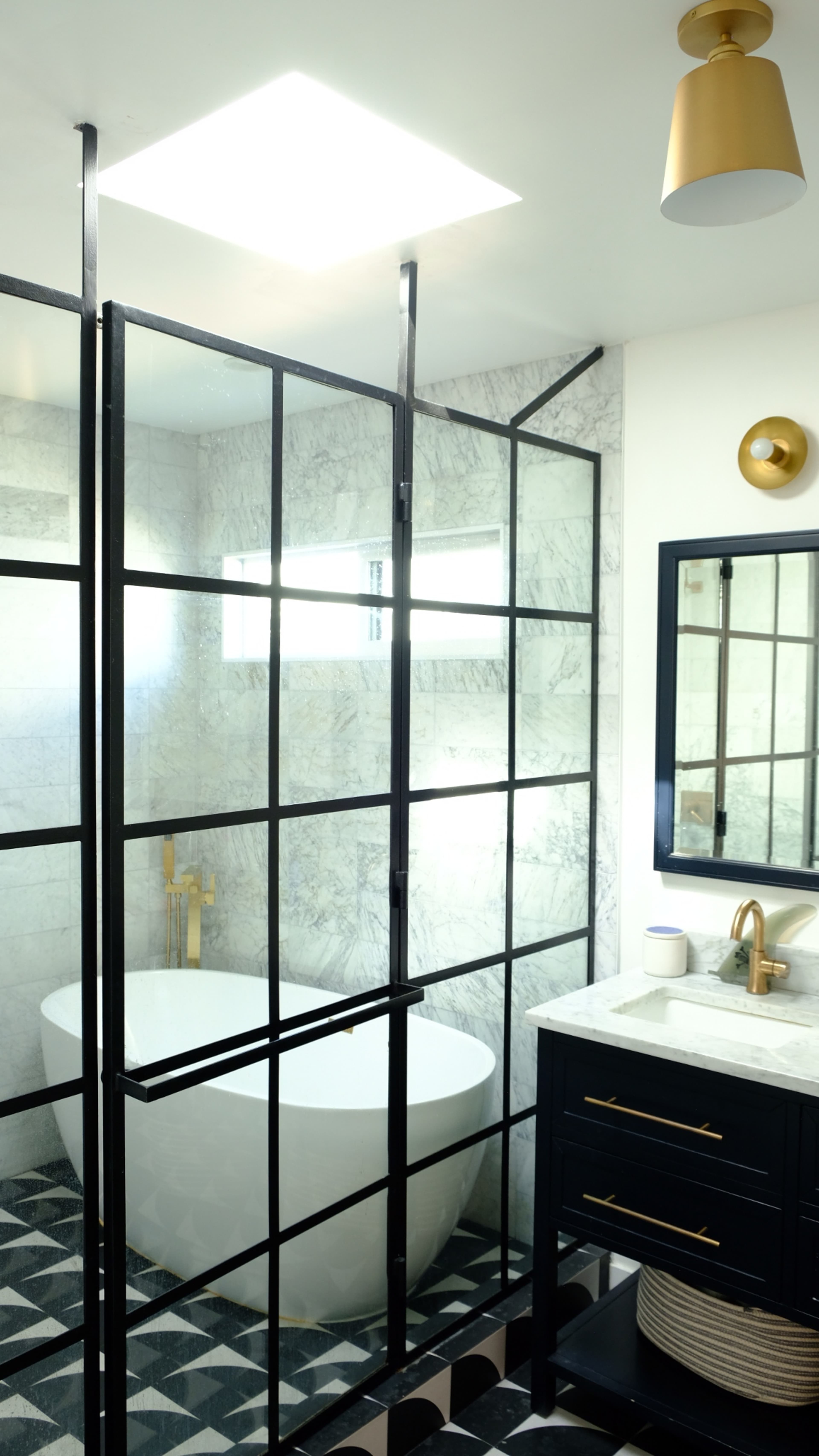 A modern bathroom featuring a freestanding tub enclosed by black framed glass partitions, with a marble wall and a vanity with a mirror.