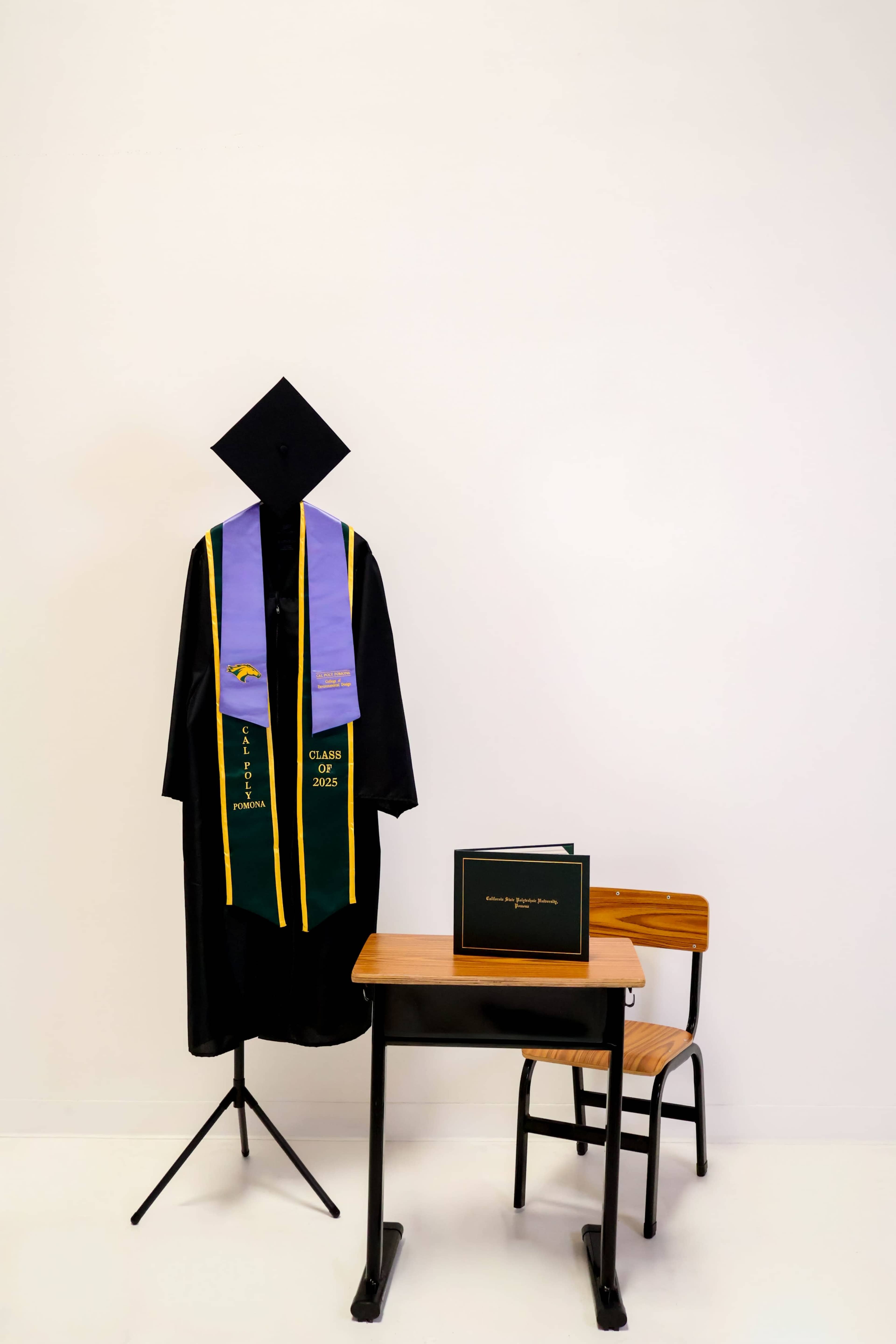 A graduation robe and cap hang on a stand next to a small desk displaying a diploma.