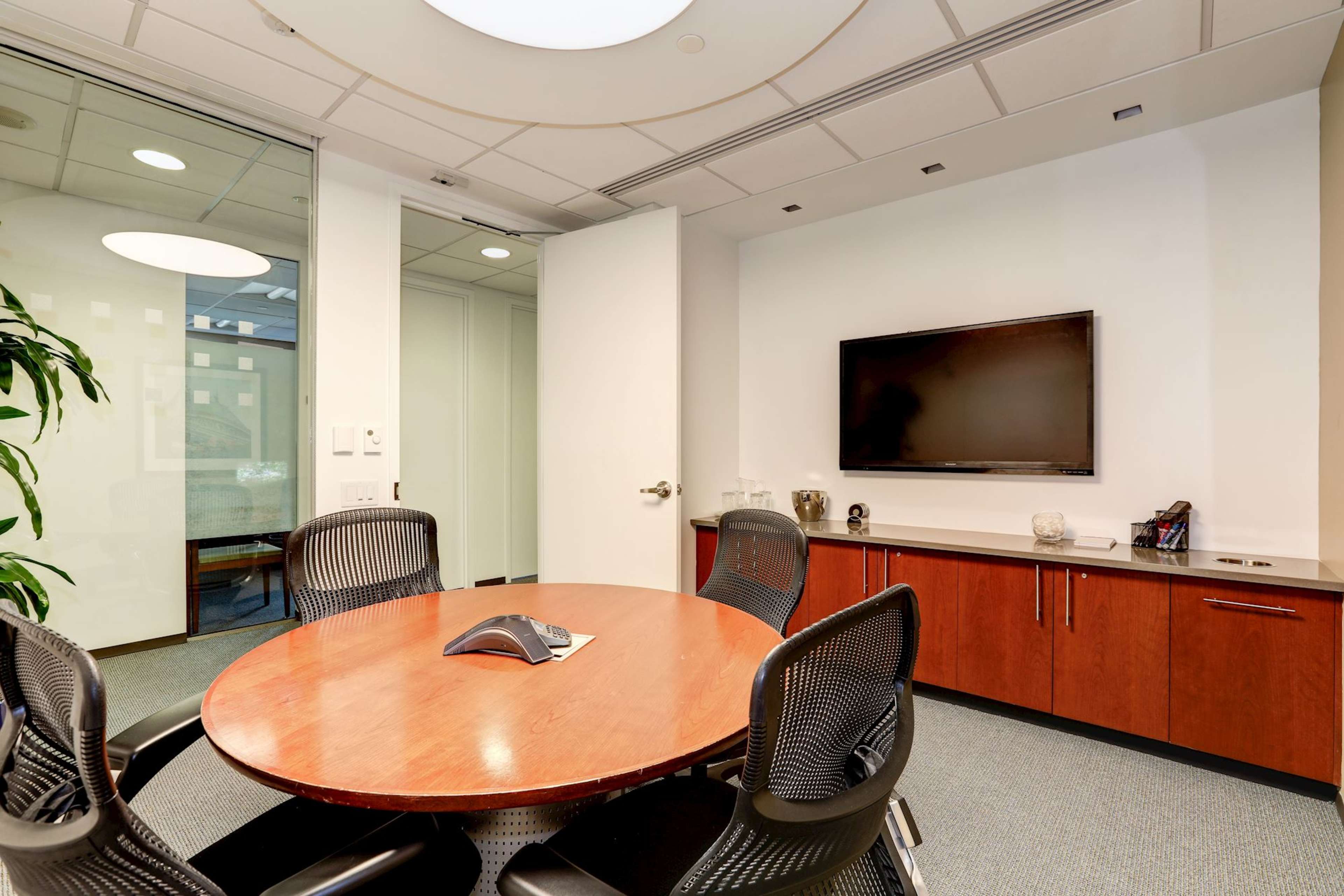 A modern conference room with a round wooden table surrounded by black mesh chairs, a television mounted on the wall, and glass partitions leading to another area.
