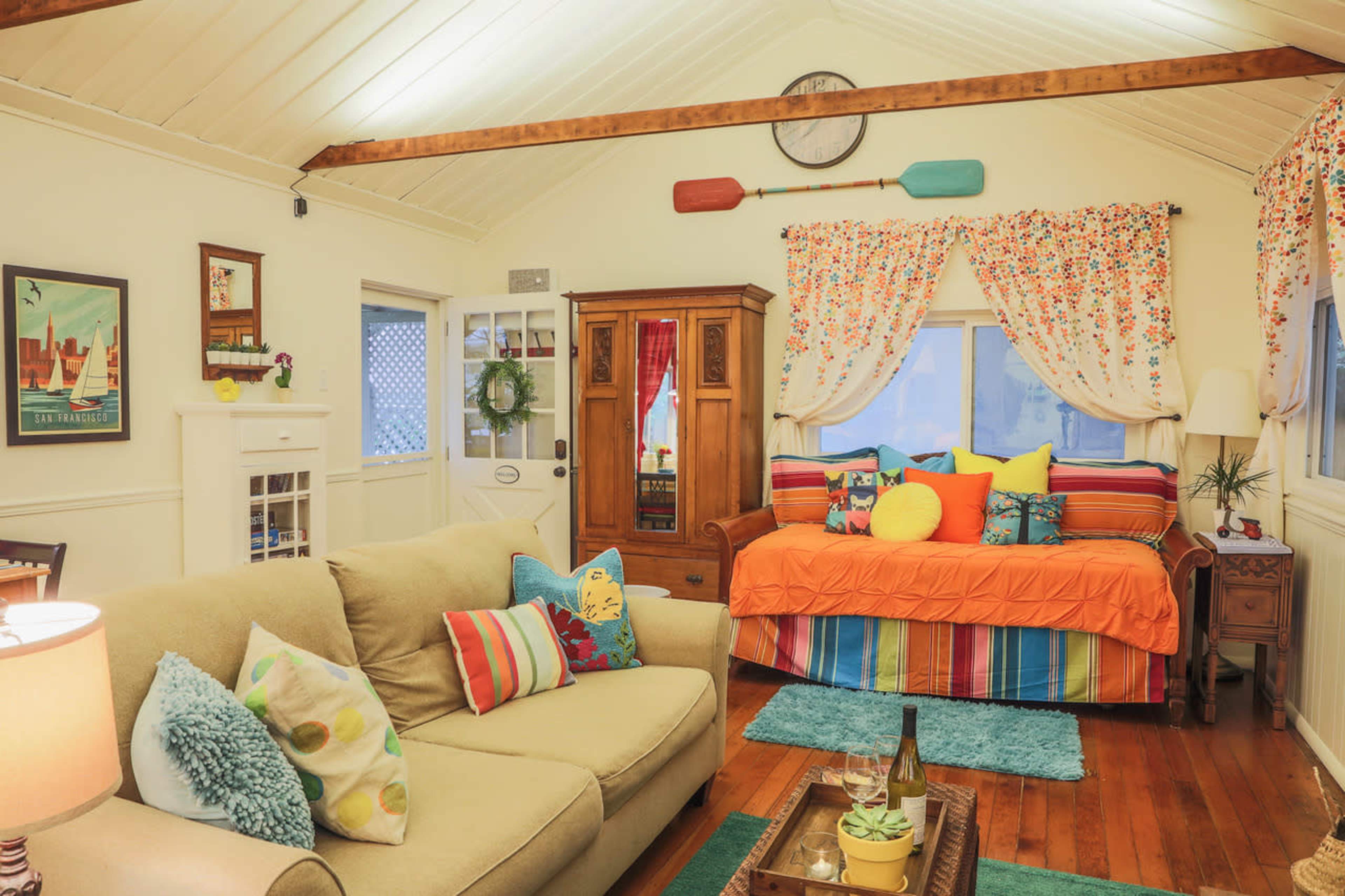A cozy living room with a couch, a daybed adorned with colorful pillows, and wooden accents, featuring a mix of floral curtains and wall art.