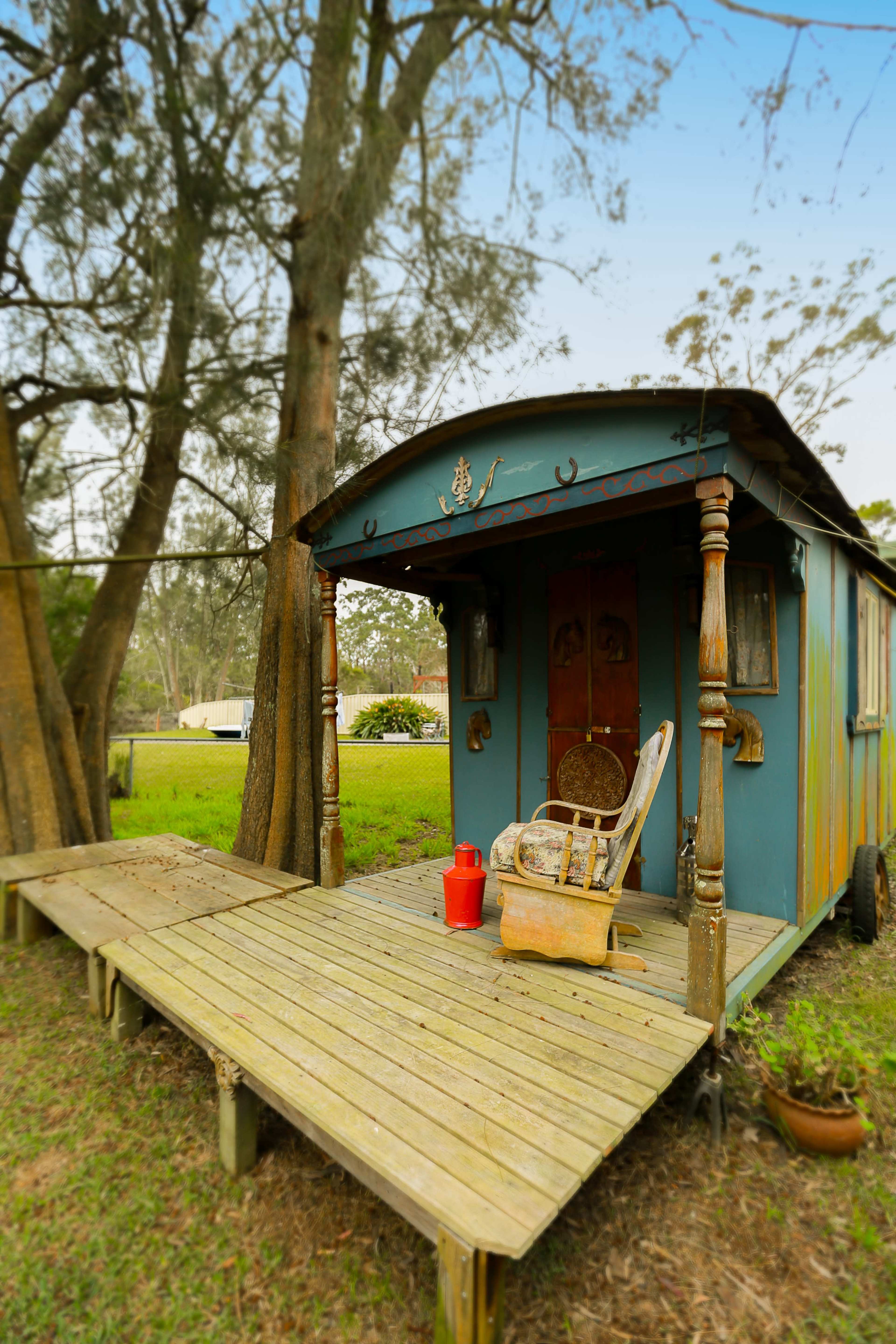 A colorful, rustic caravan sits on a wooden deck surrounded by trees in a grassy area.