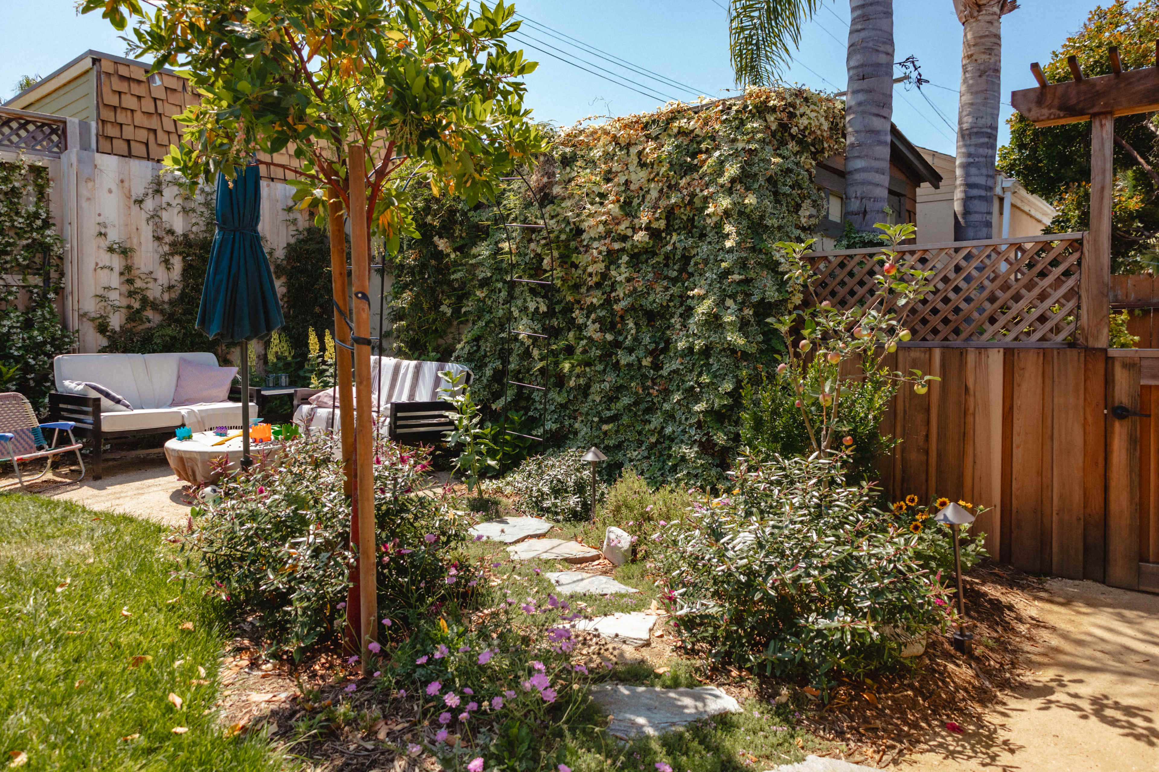 The image shows a well-maintained backyard garden featuring a stone pathway, colorful flowers, a small seating area, and a tall green shrub wall.