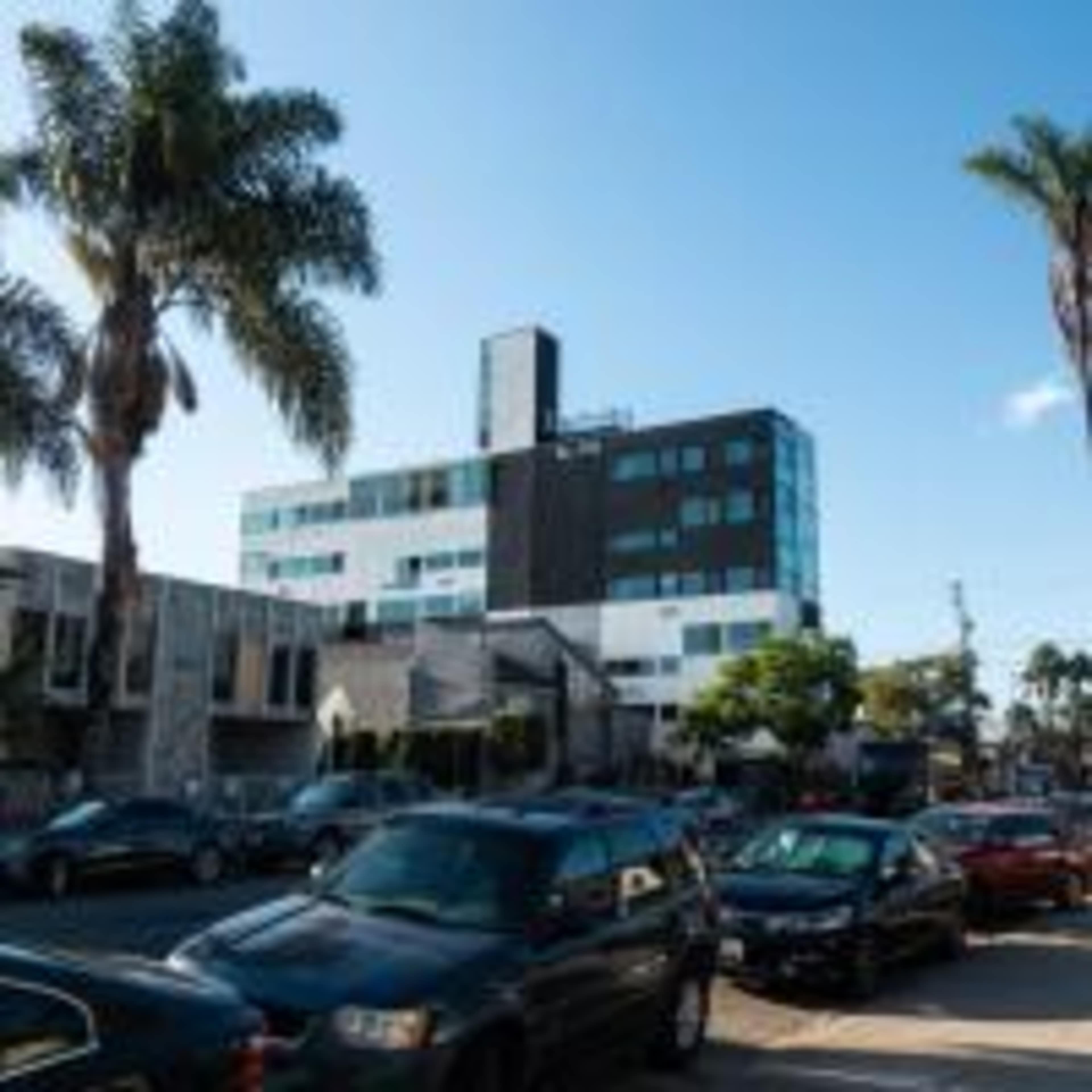 The image shows a modern building with a mix of black and white facades, situated alongside a street lined with parked cars and palm trees.
