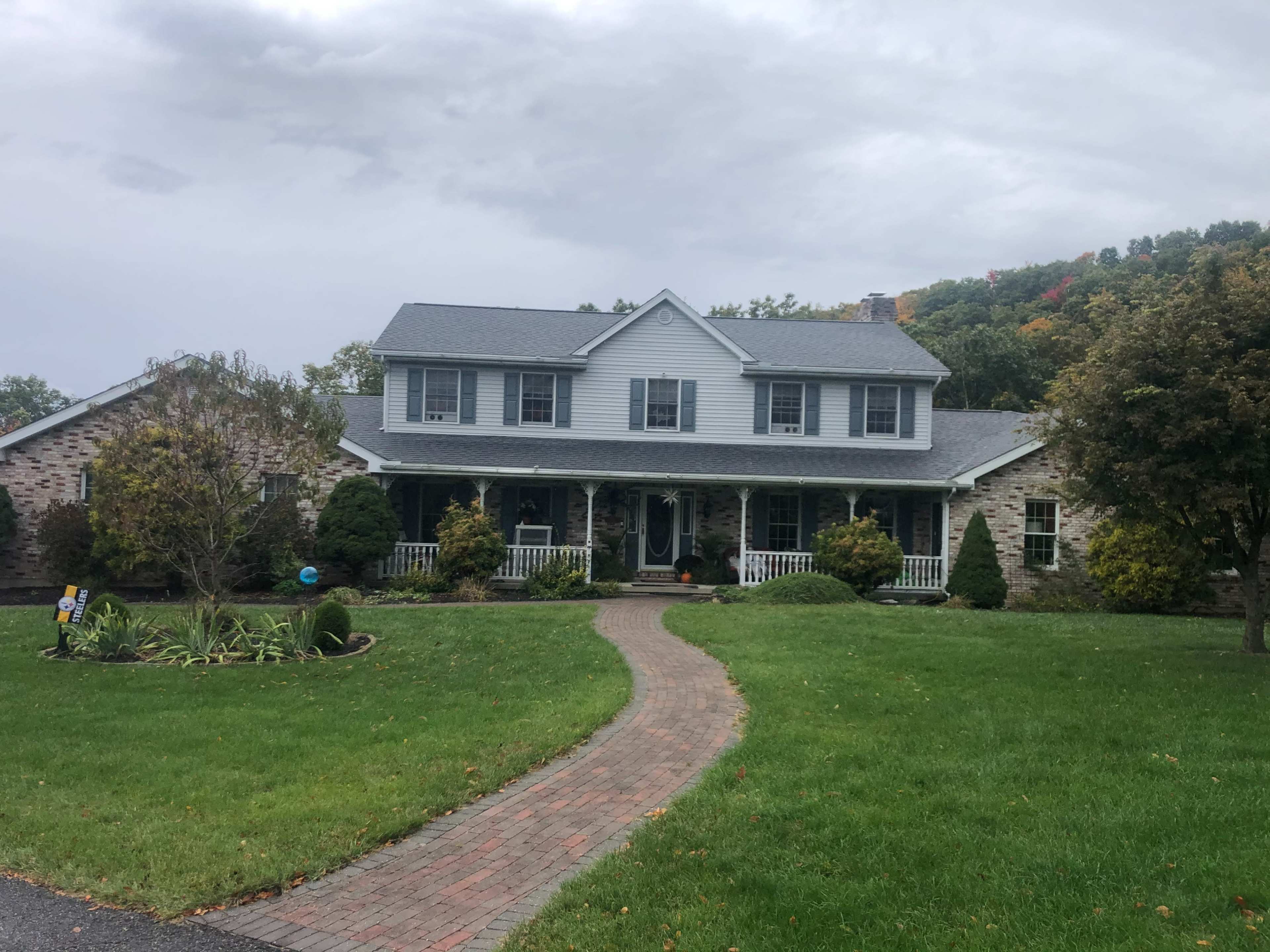 A two-story gray house with a front porch is set on a landscaped lawn featuring a curved brick pathway.
