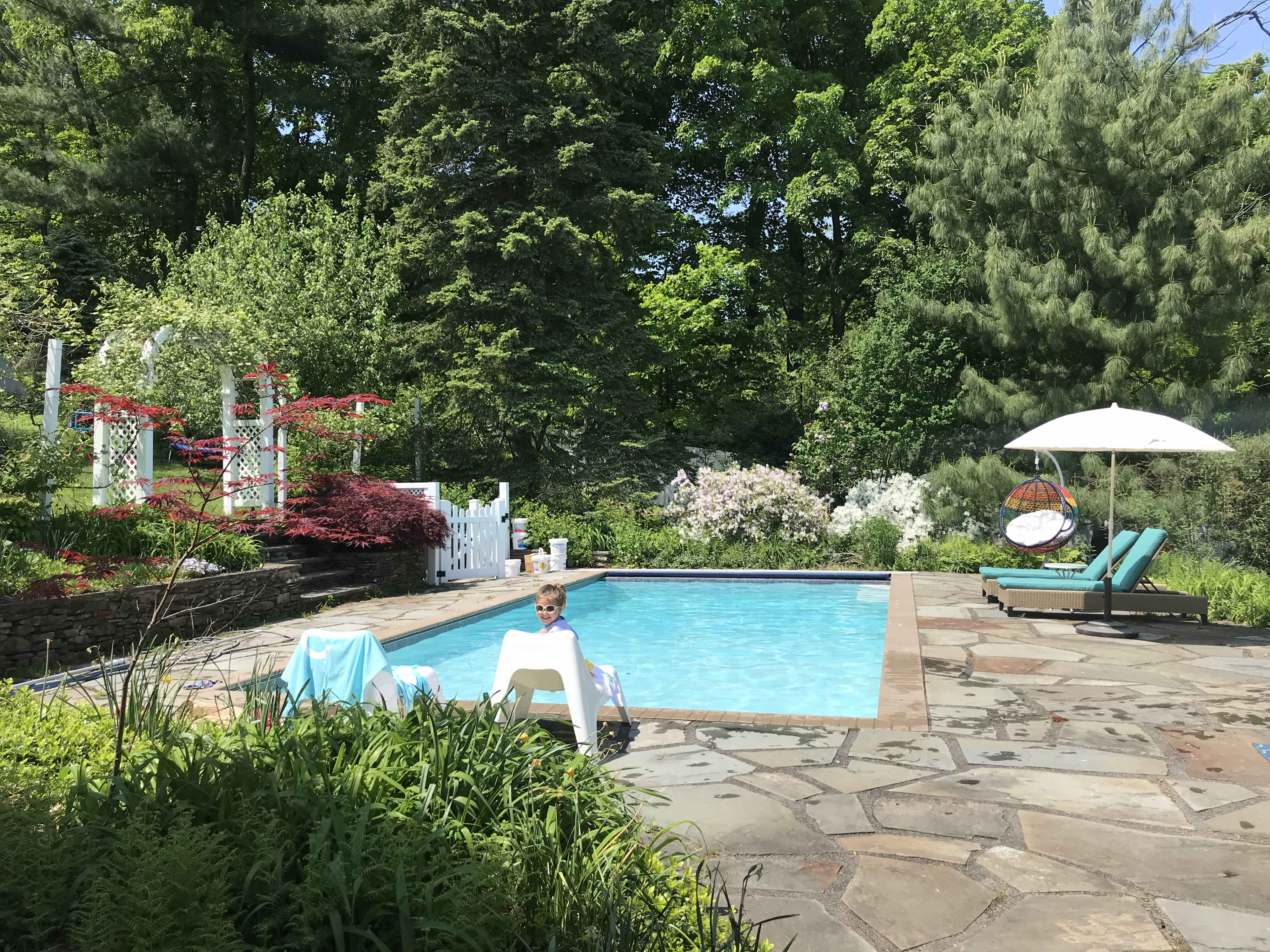 A child sits on a white lounge chair beside a swimming pool surrounded by greenery and colorful flowers.