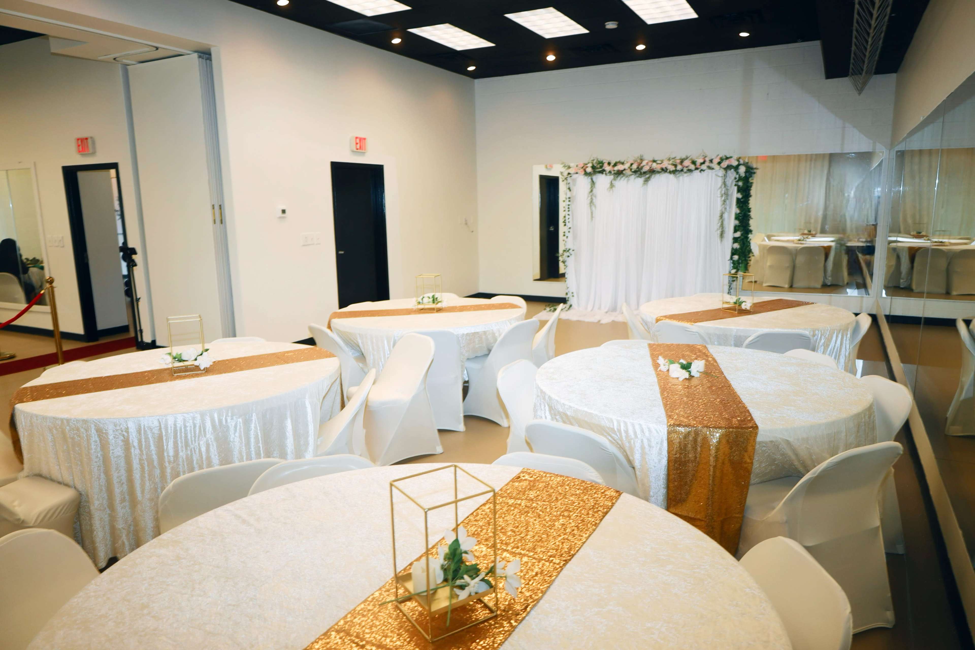 A decorated event space with several round tables covered in white tablecloths, gold runners, and a backdrop with greenery and drapery at one end.