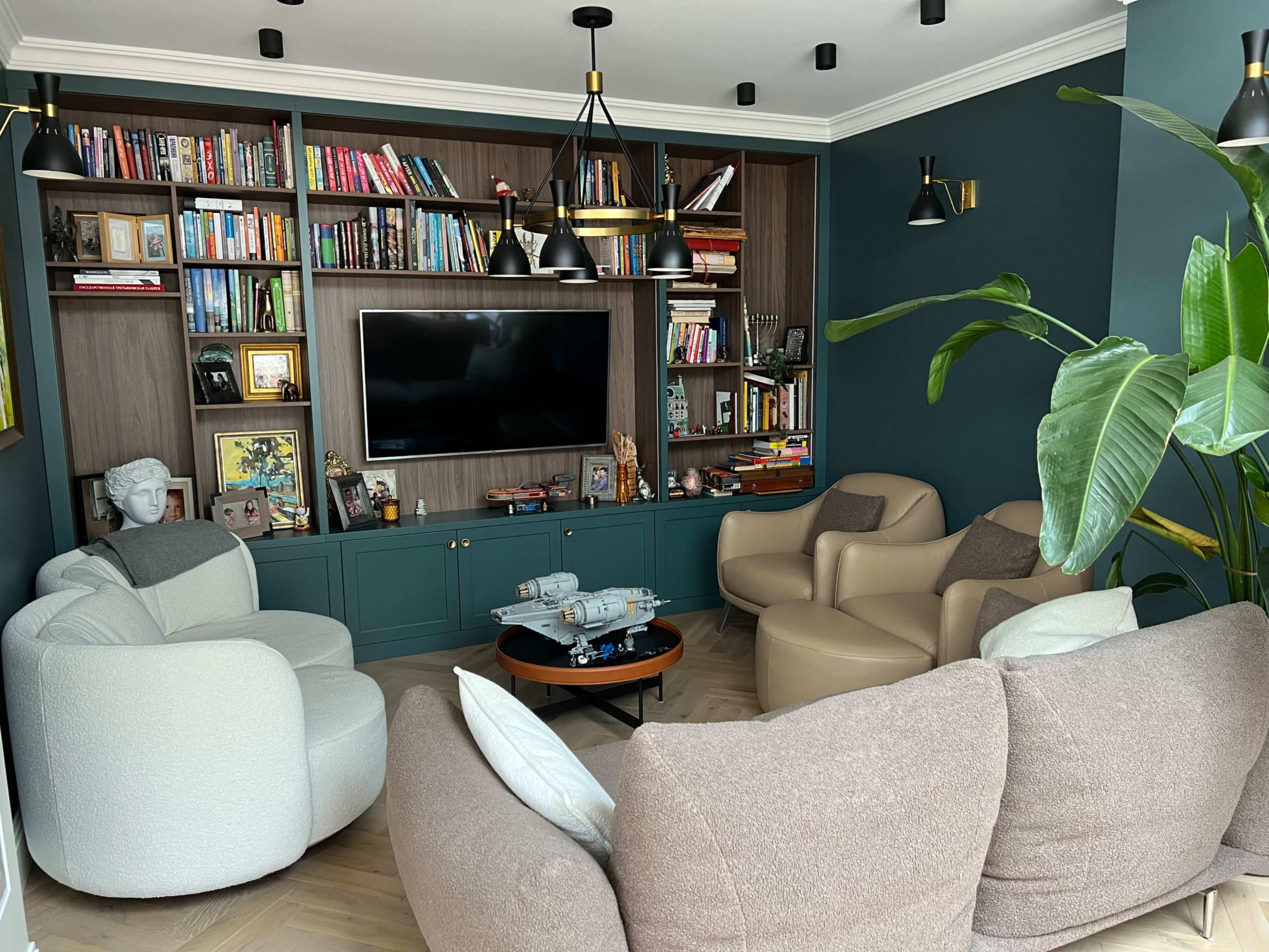 A cozy living room features a wall of bookshelves, a television, and two distinct seating areas with modern armchairs and a round coffee table.