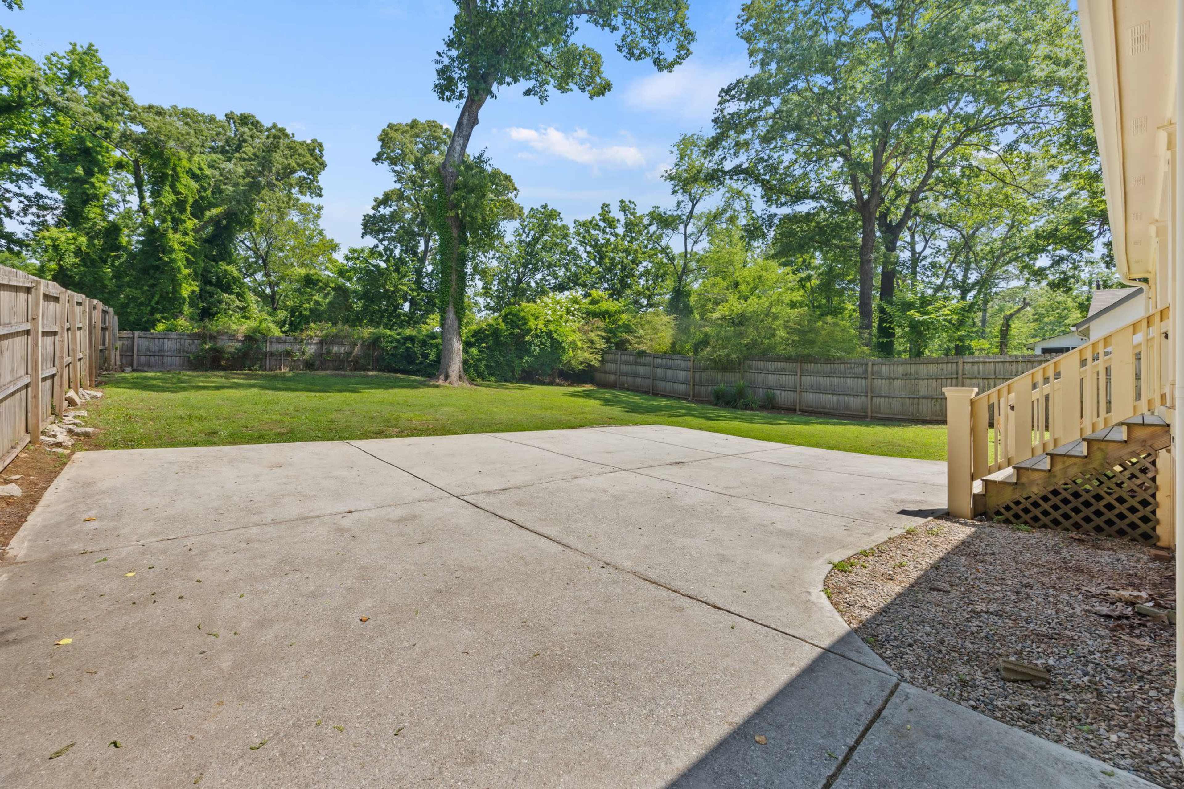 The image shows a spacious, flat backyard enclosed by a wooden fence, with a concrete patio leading to the house.