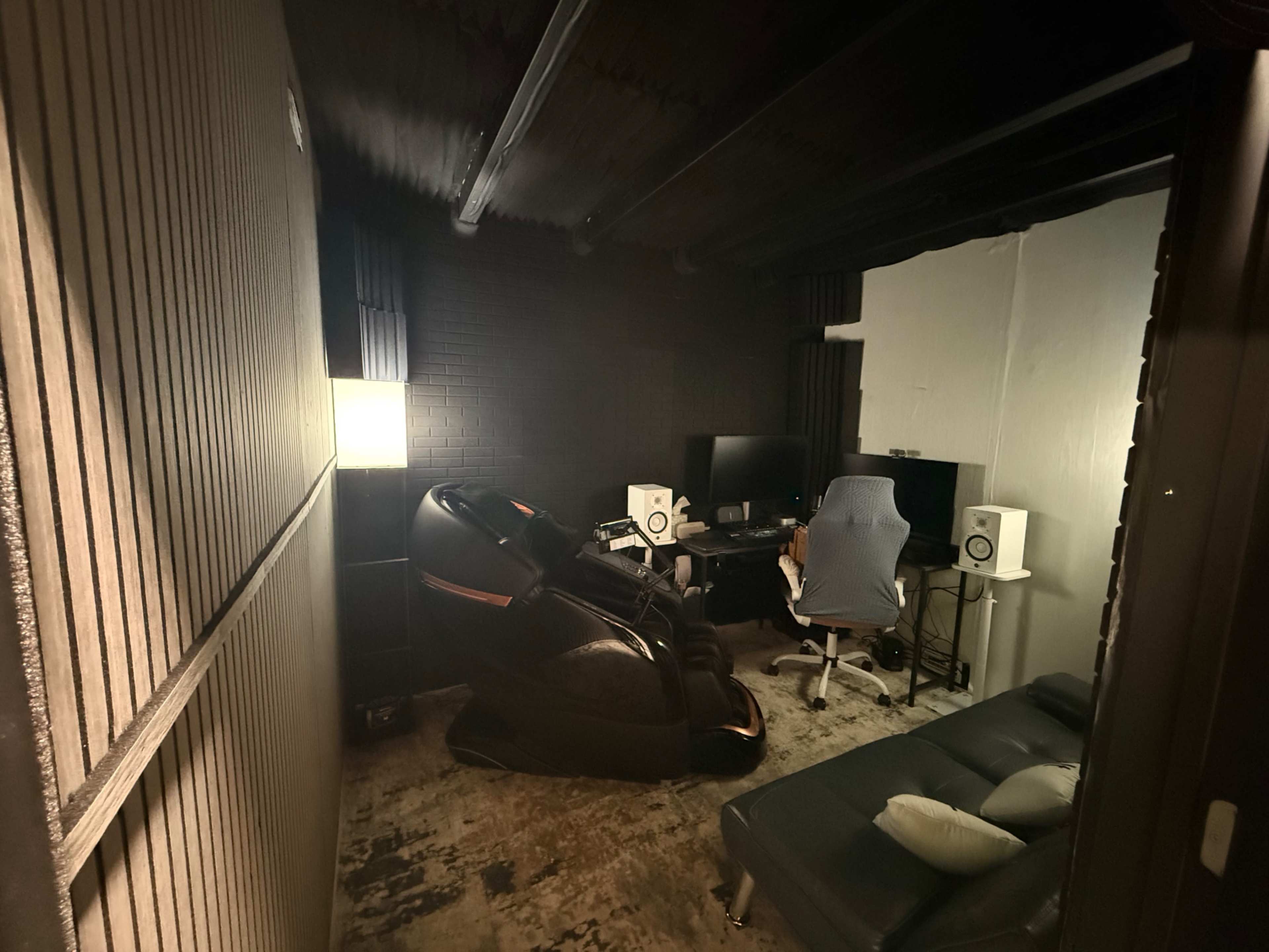 The image shows a small, dimly lit room furnished with a massage chair, a computer desk with monitors, and speakers against the walls.