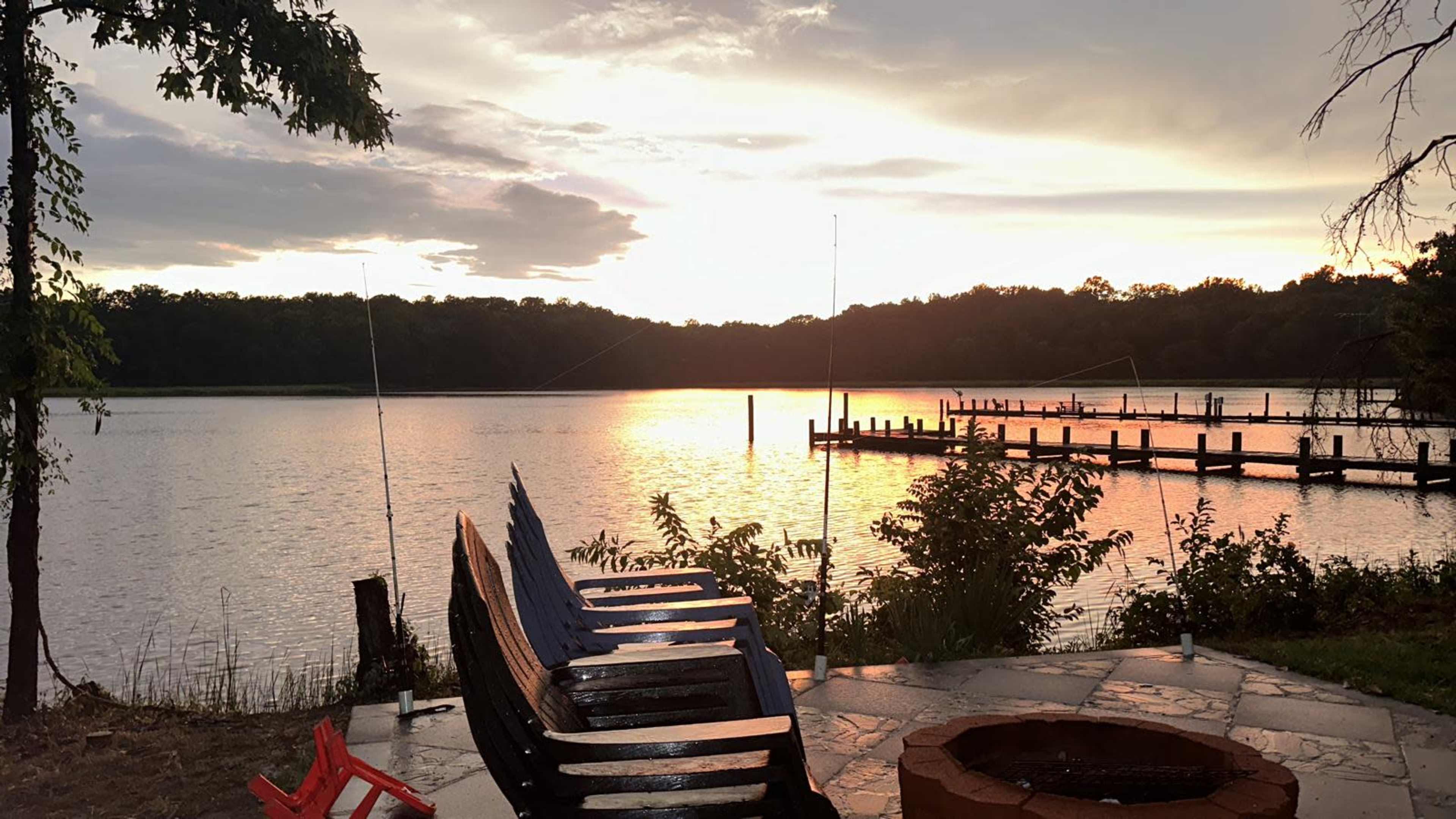 A row of wooden chairs overlooks a lake at sunset, with a dock and fishing poles visible in the background.