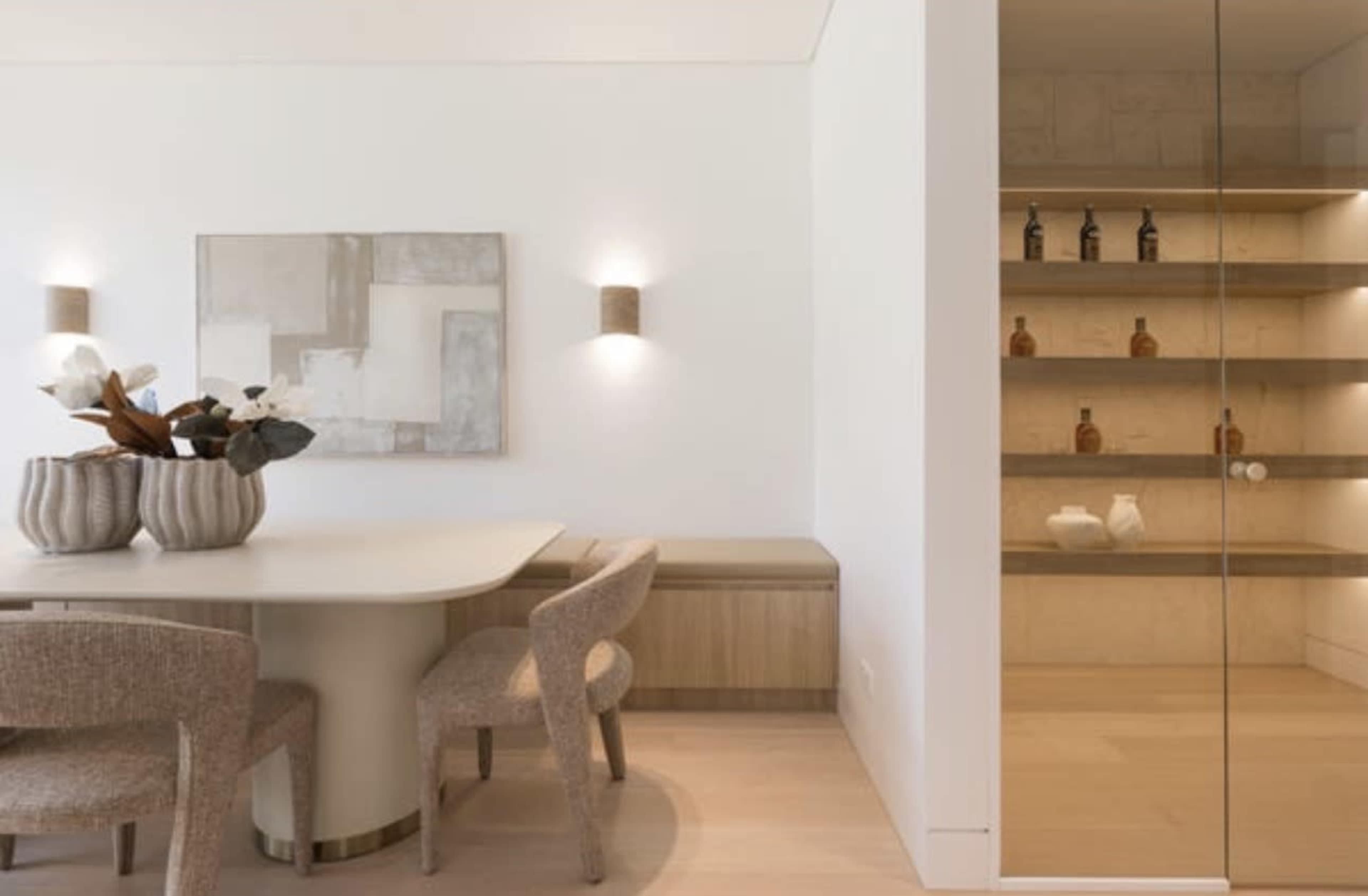 The image shows a modern dining area featuring a round table surrounded by four beige chairs, with a decorative wall art piece and an adjacent glass-fronted shelving unit displaying various bottles and items.