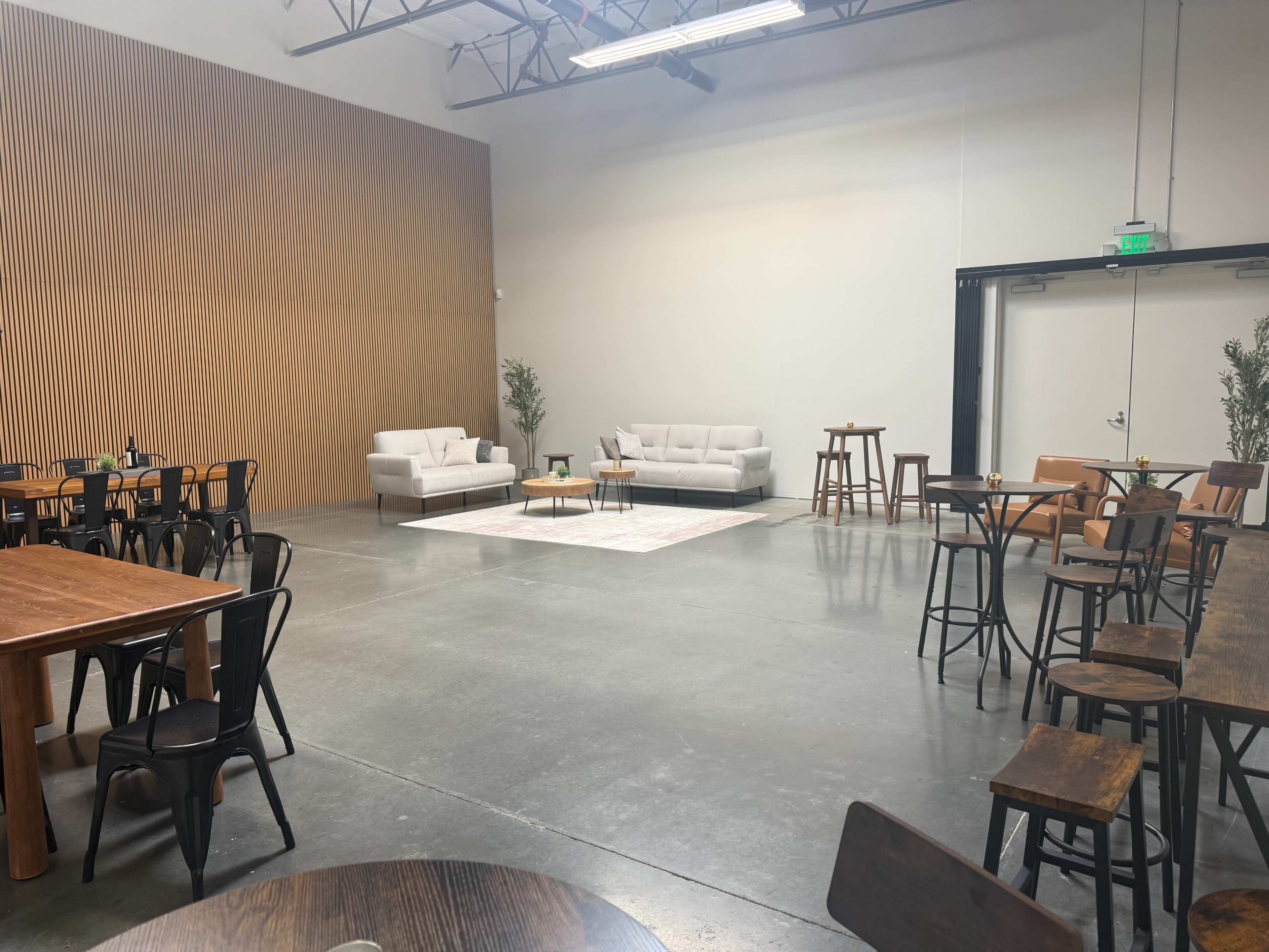 The image shows a spacious, modern venue featuring a mix of seating arrangements, including tables and chairs, with a neutral color palette and wooden accents.