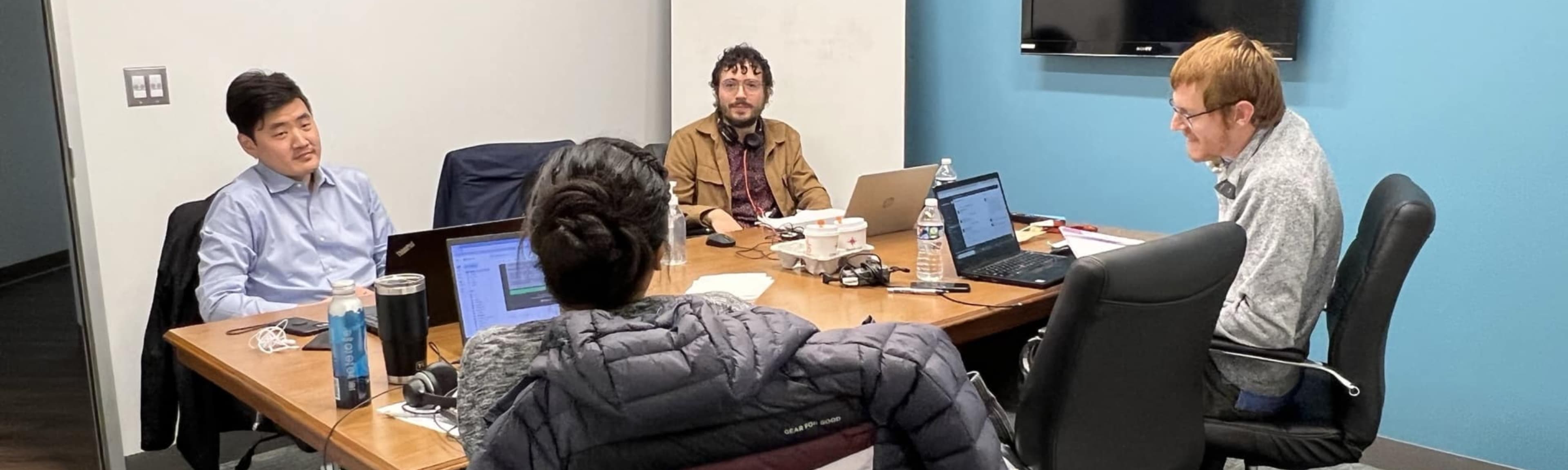 A group of four people sit around a conference table, engaged in discussion, with laptops and documents in front of them.