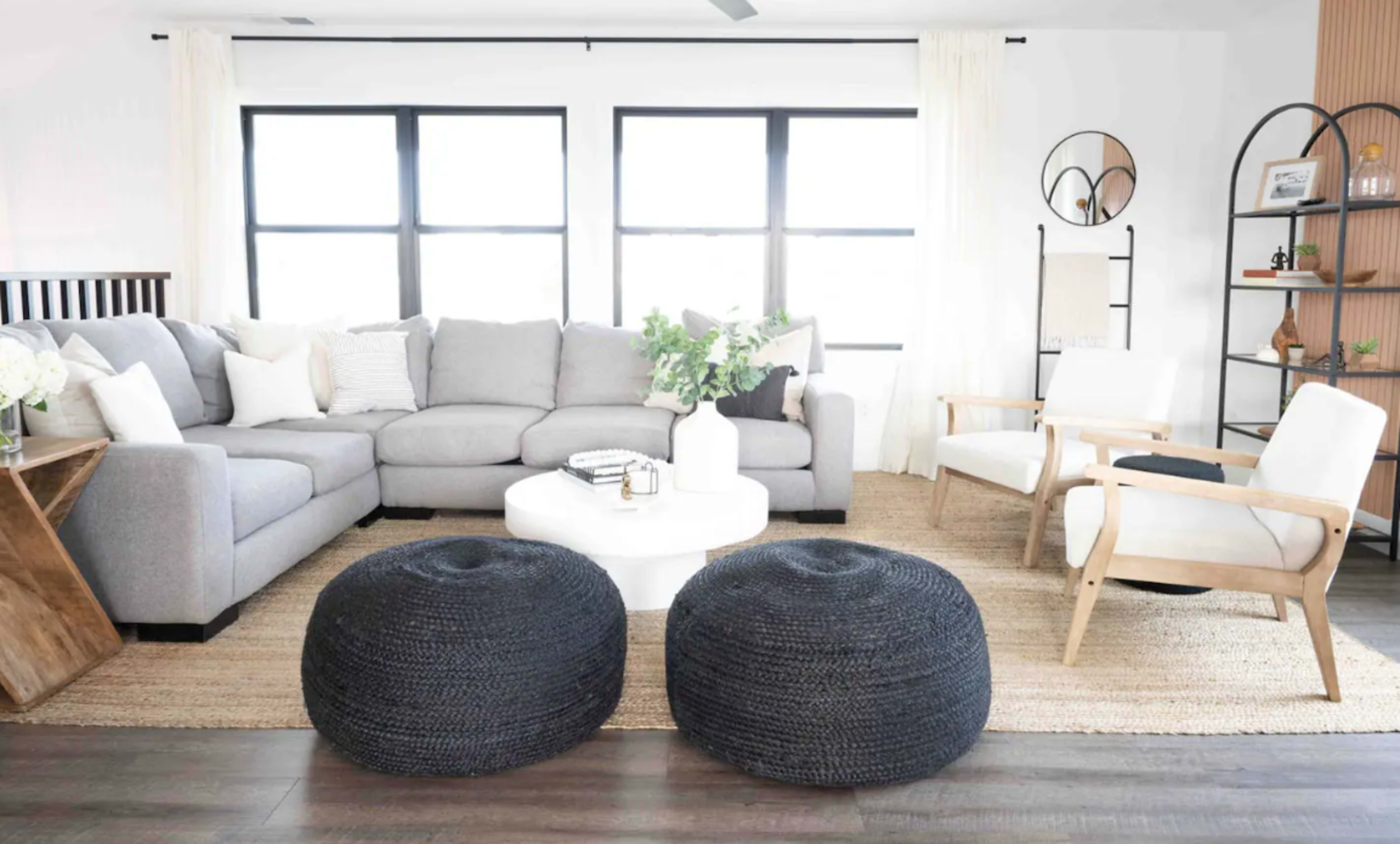A modern living room features a gray sectional sofa, a round coffee table, two black poufs, and an accent chair, all arranged on a textured rug.