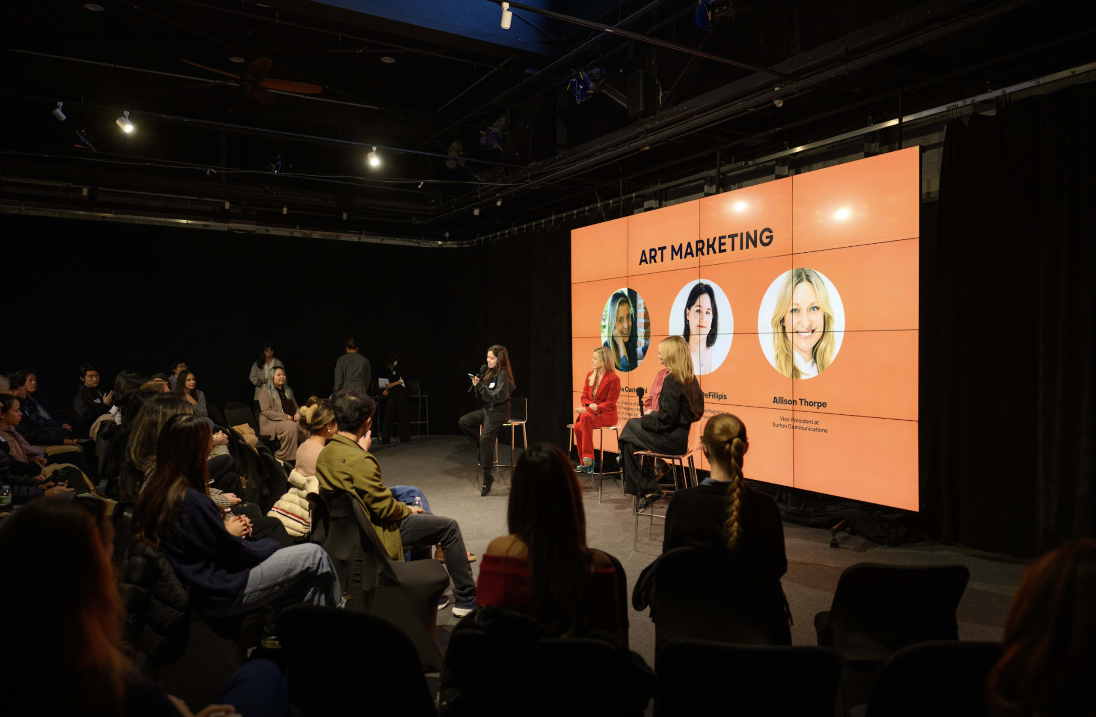 A panel discussion on art marketing is taking place in a dark room with an audience seated in front of a large screen displaying the title and images of the speakers.