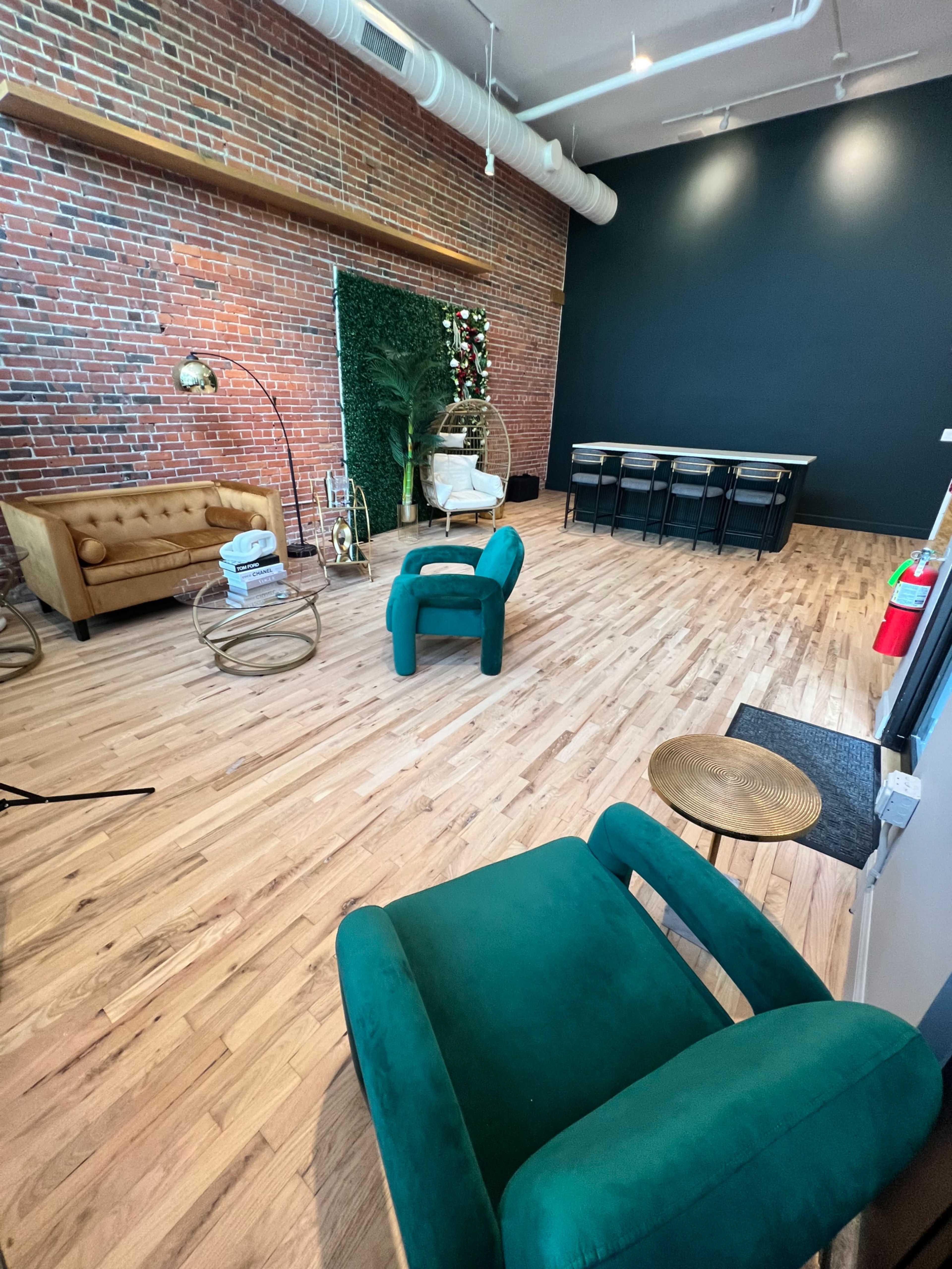 A spacious interior with hardwood flooring, featuring a green chair, a beige sofa, a dining table with chairs, and a decorative plant against a brick wall.