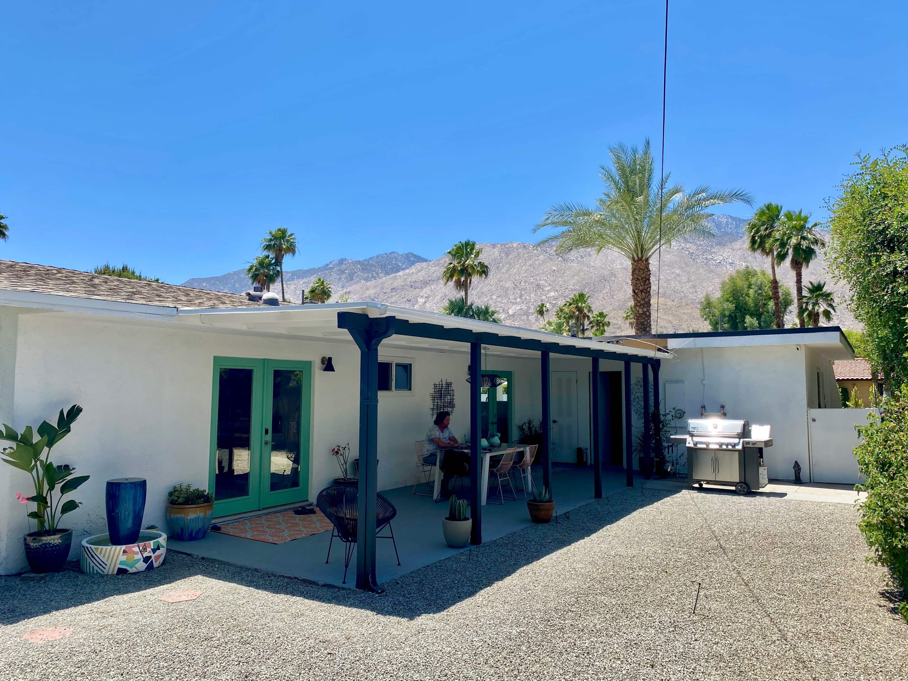 A white house with a patio surrounded by palm trees and mountains in the background.