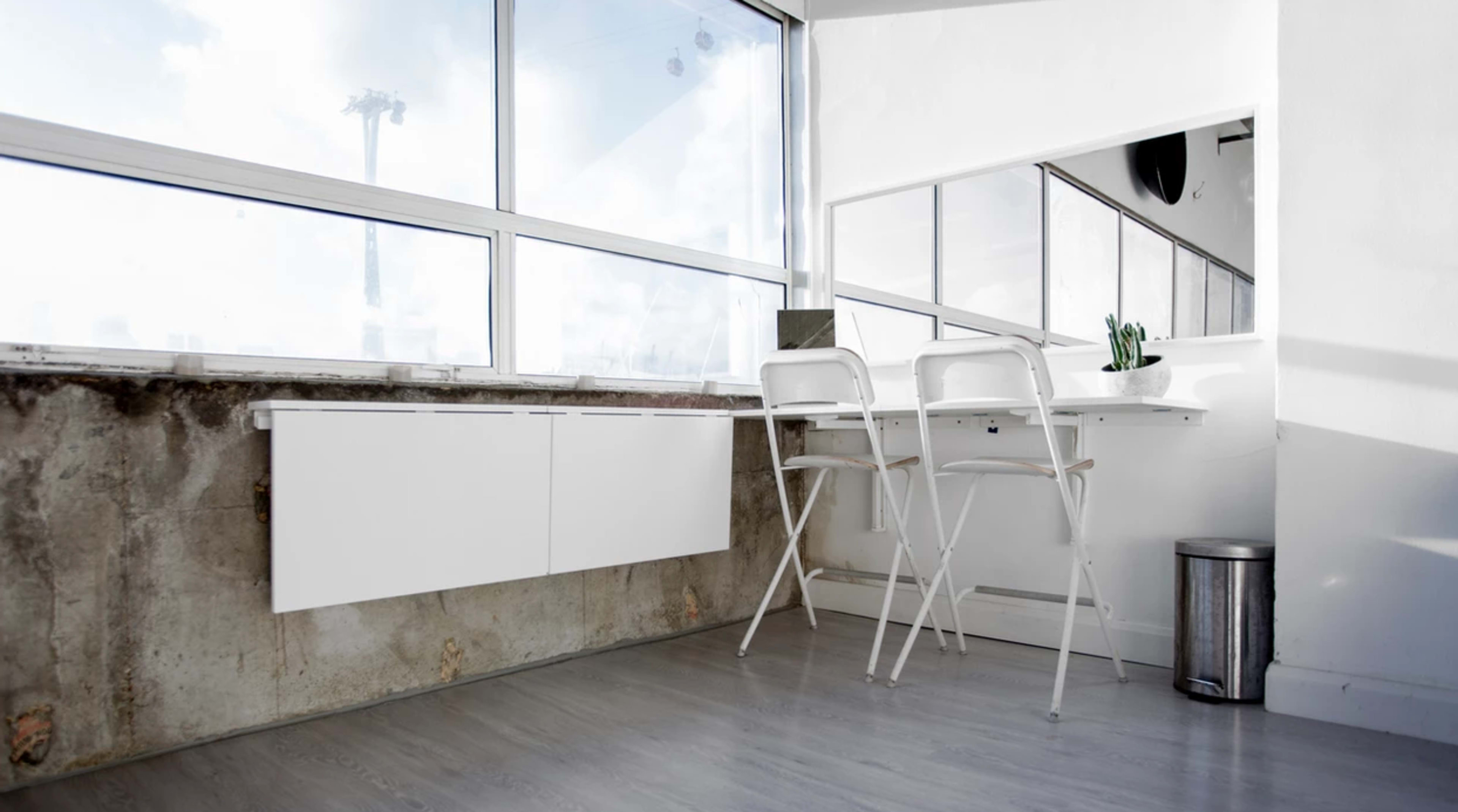 A bright workspace features a white table and two chairs beside large windows with a concrete wall.