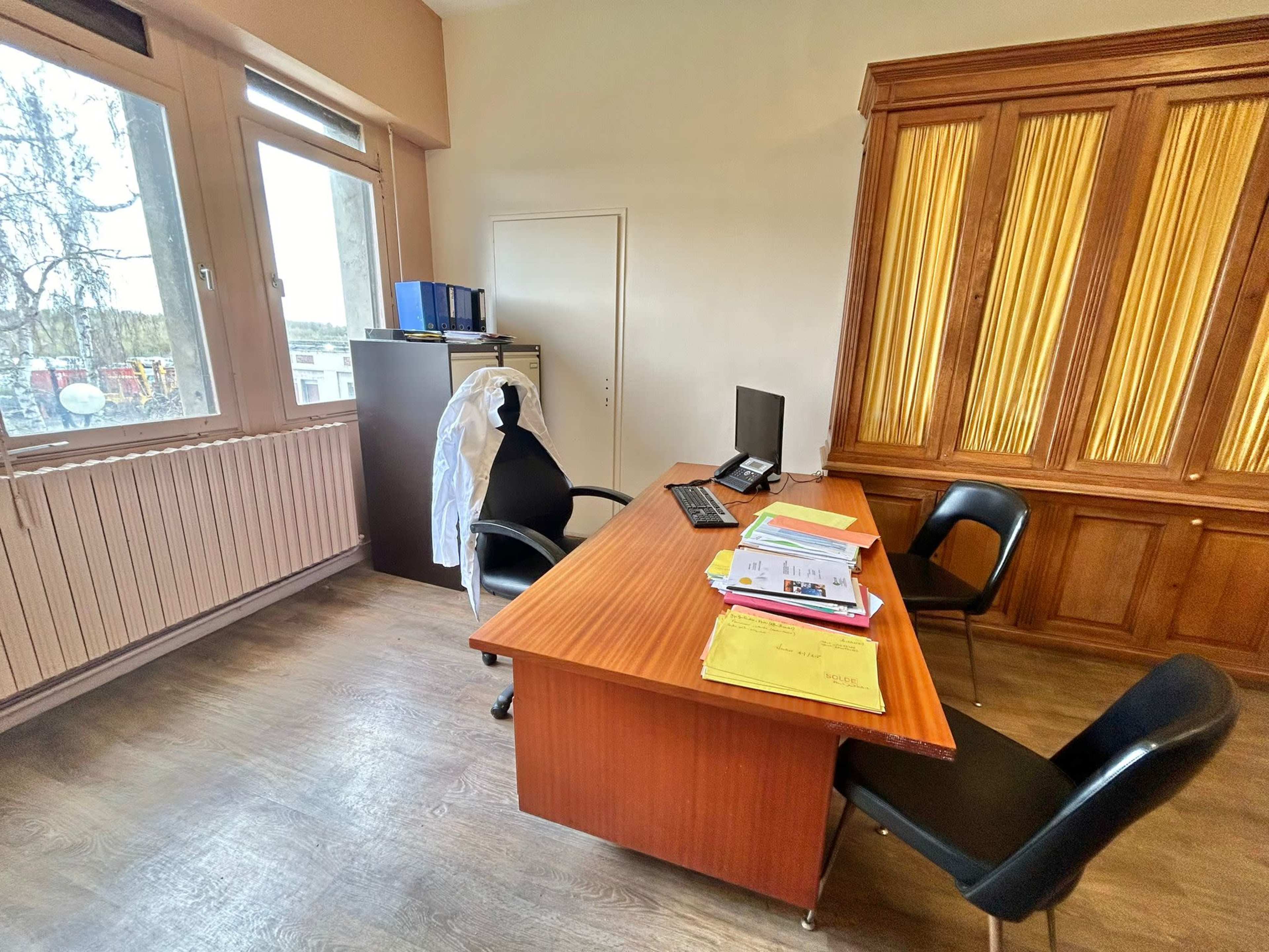 A well-organized office with a wooden desk, a computer, and a stack of papers, set against a backdrop of large windows and wooden cabinetry.