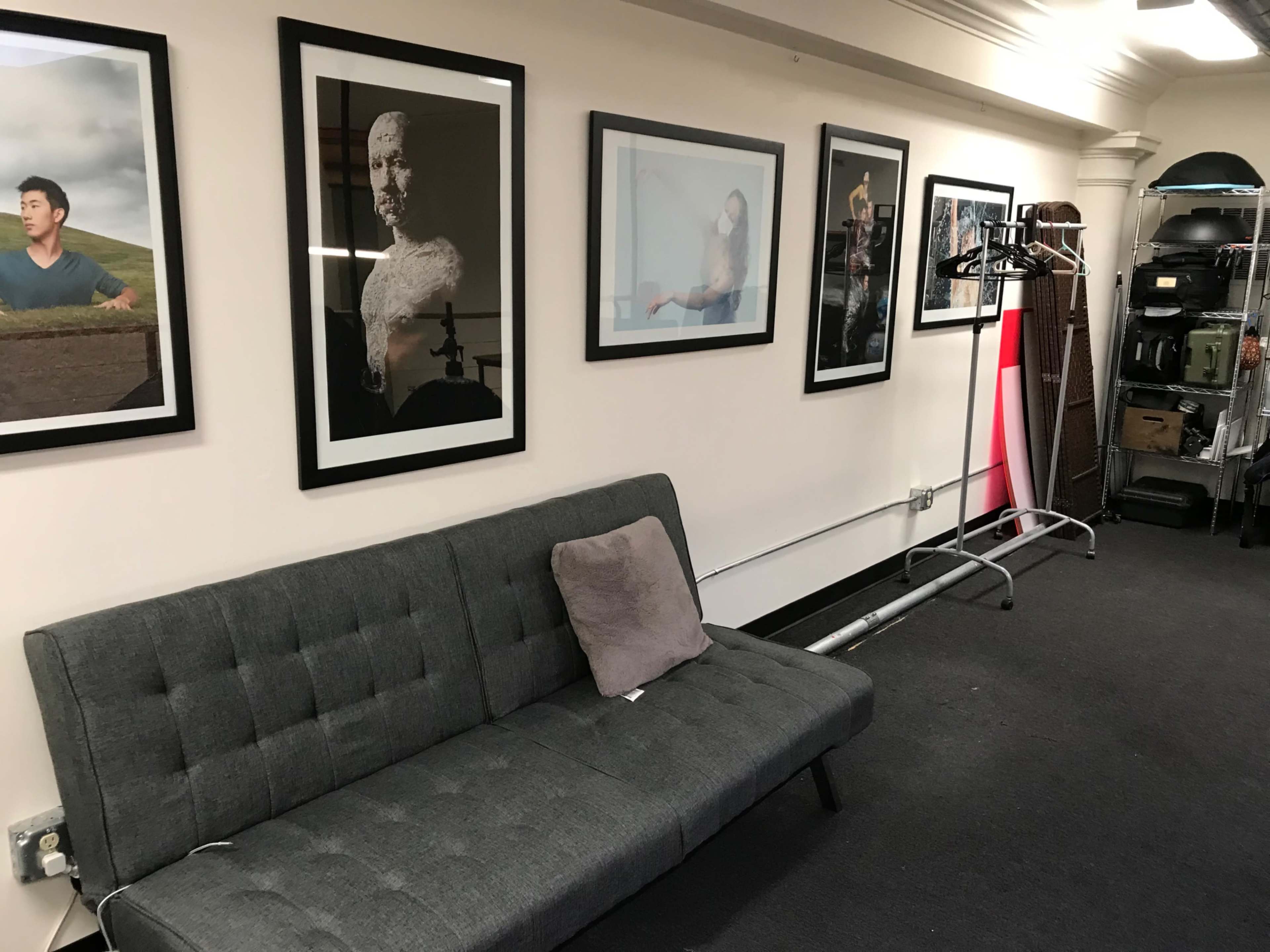 A gray sofa sits against a wall adorned with framed black-and-white photographs, with a garment rack and various items visible in a cluttered area to the side.