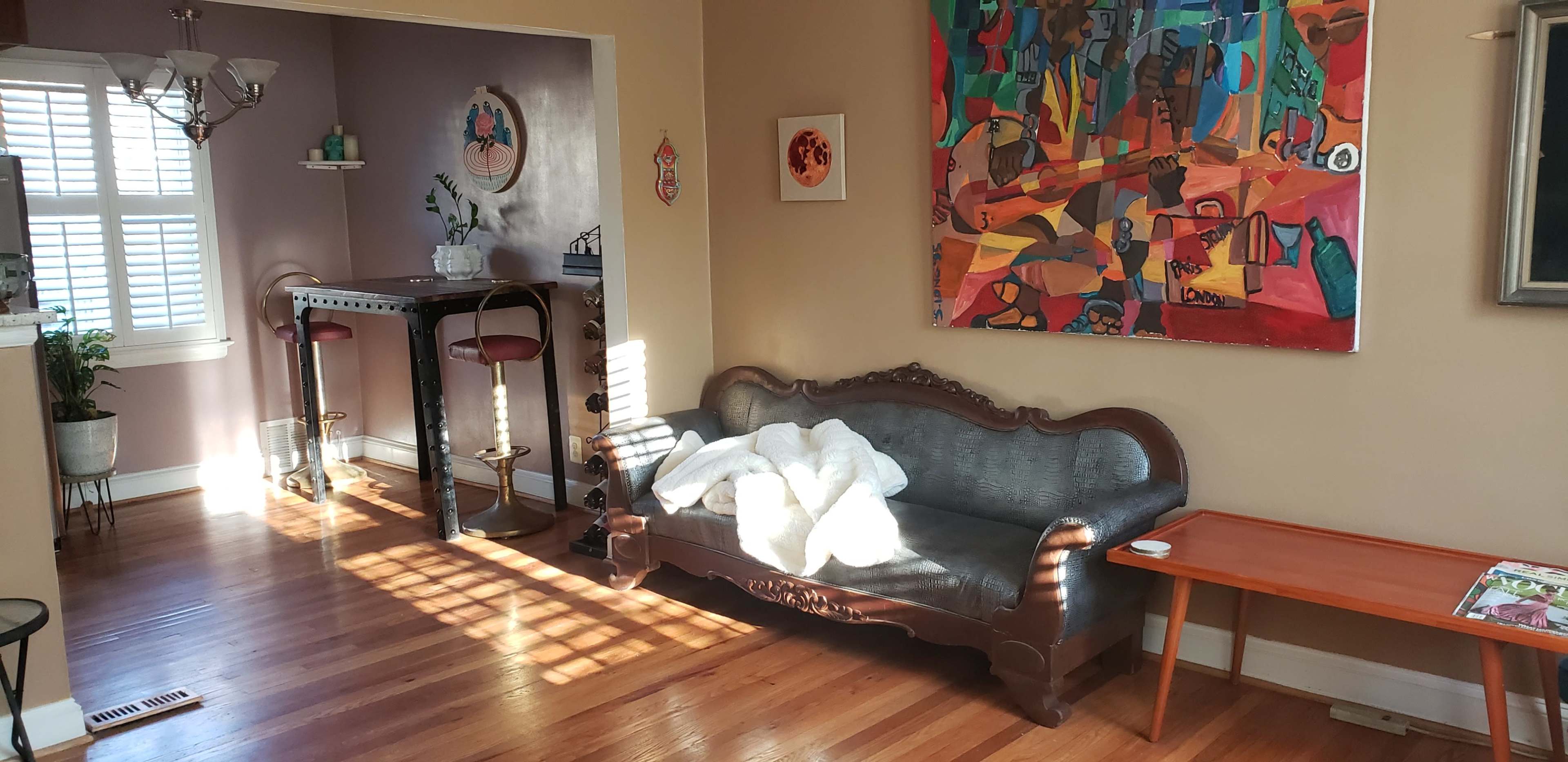 A living room features a dark wooden sofa draped with a white blanket, a small table, and a bar area with stools, alongside a colorful abstract painting on the wall.