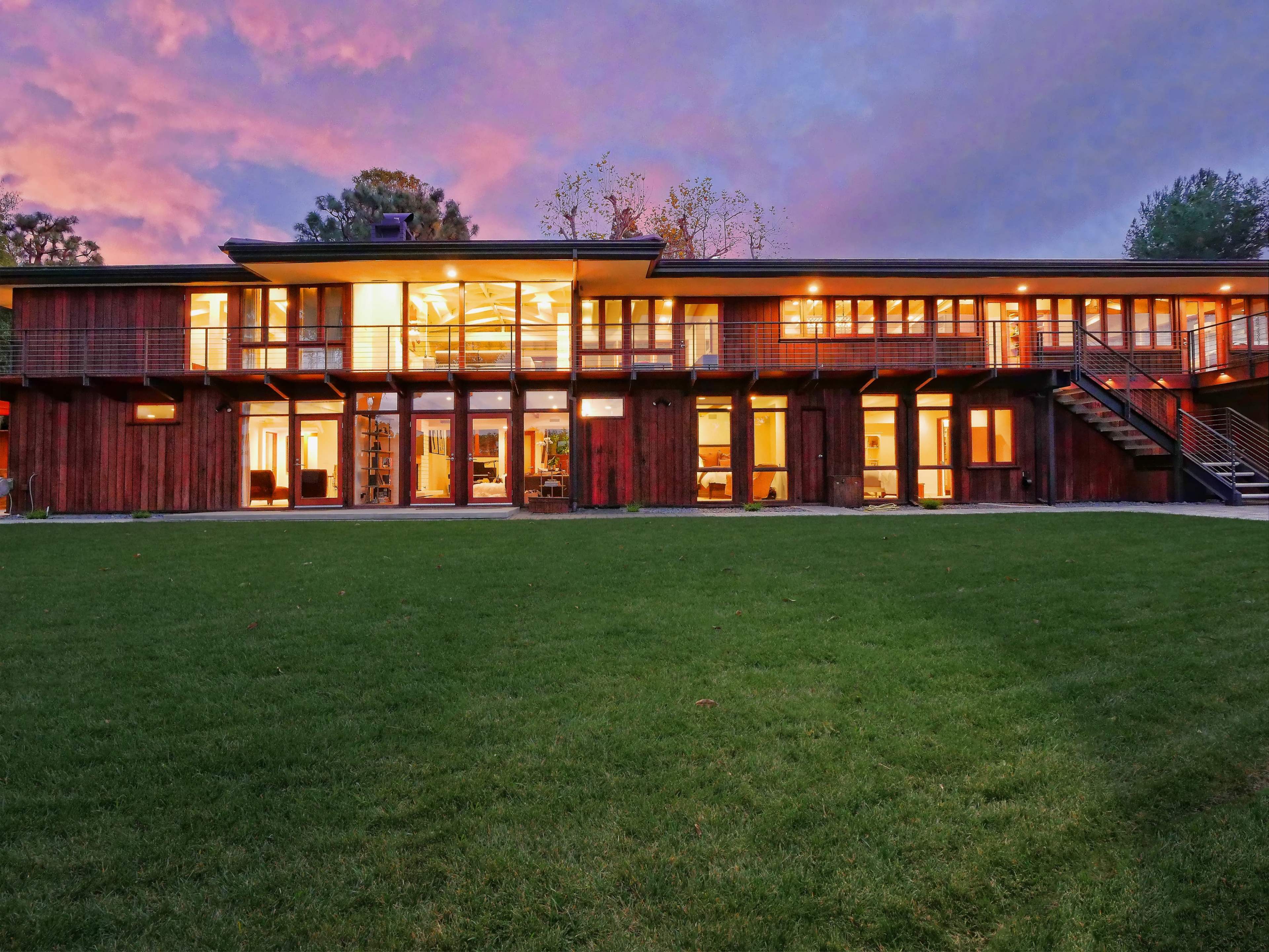 The image shows a modern two-story house with large glass windows and a wooden exterior, set against a colorful sunset sky and a green lawn.