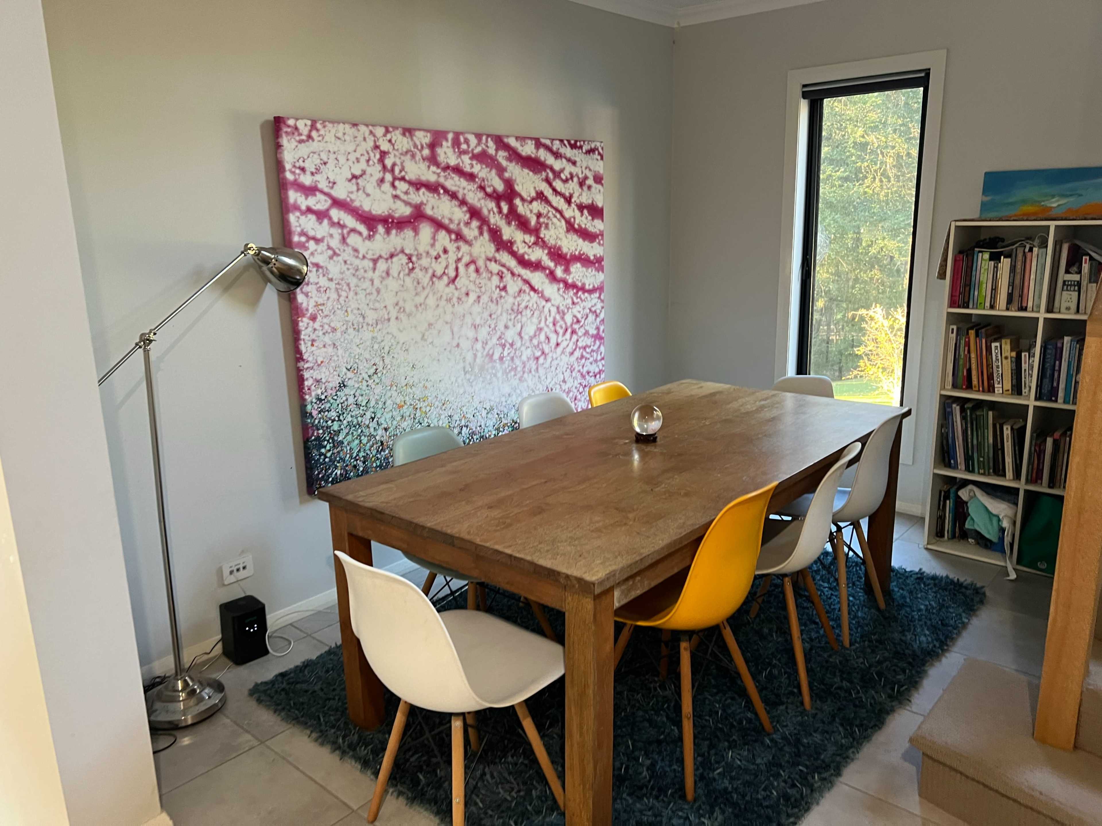 A dining area features a large wooden table surrounded by colorful chairs, with a lamp and a bookshelf in the background.