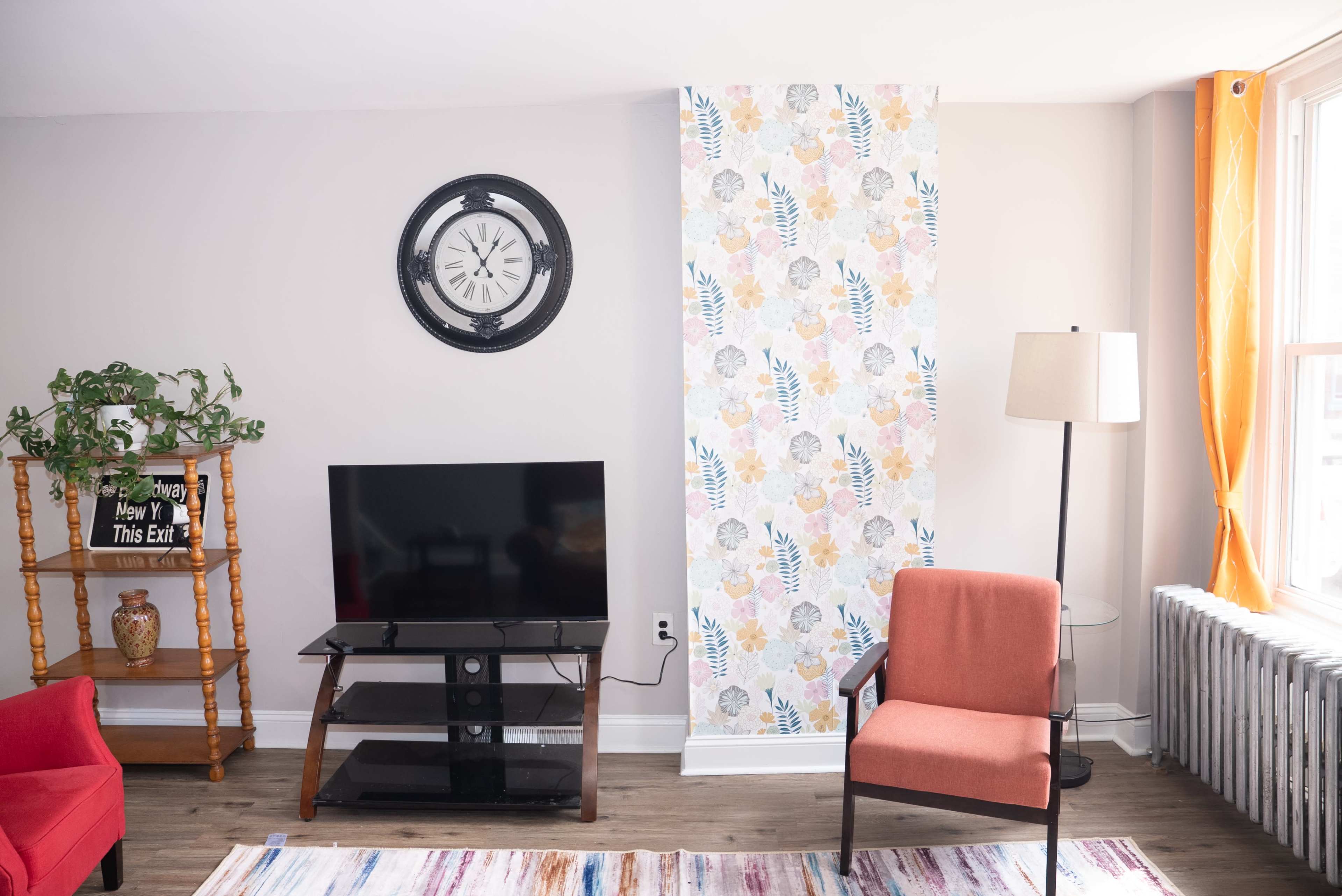 The image shows a living room with a wall clock, a television stand, an orange chair, and a patterned feature wall.