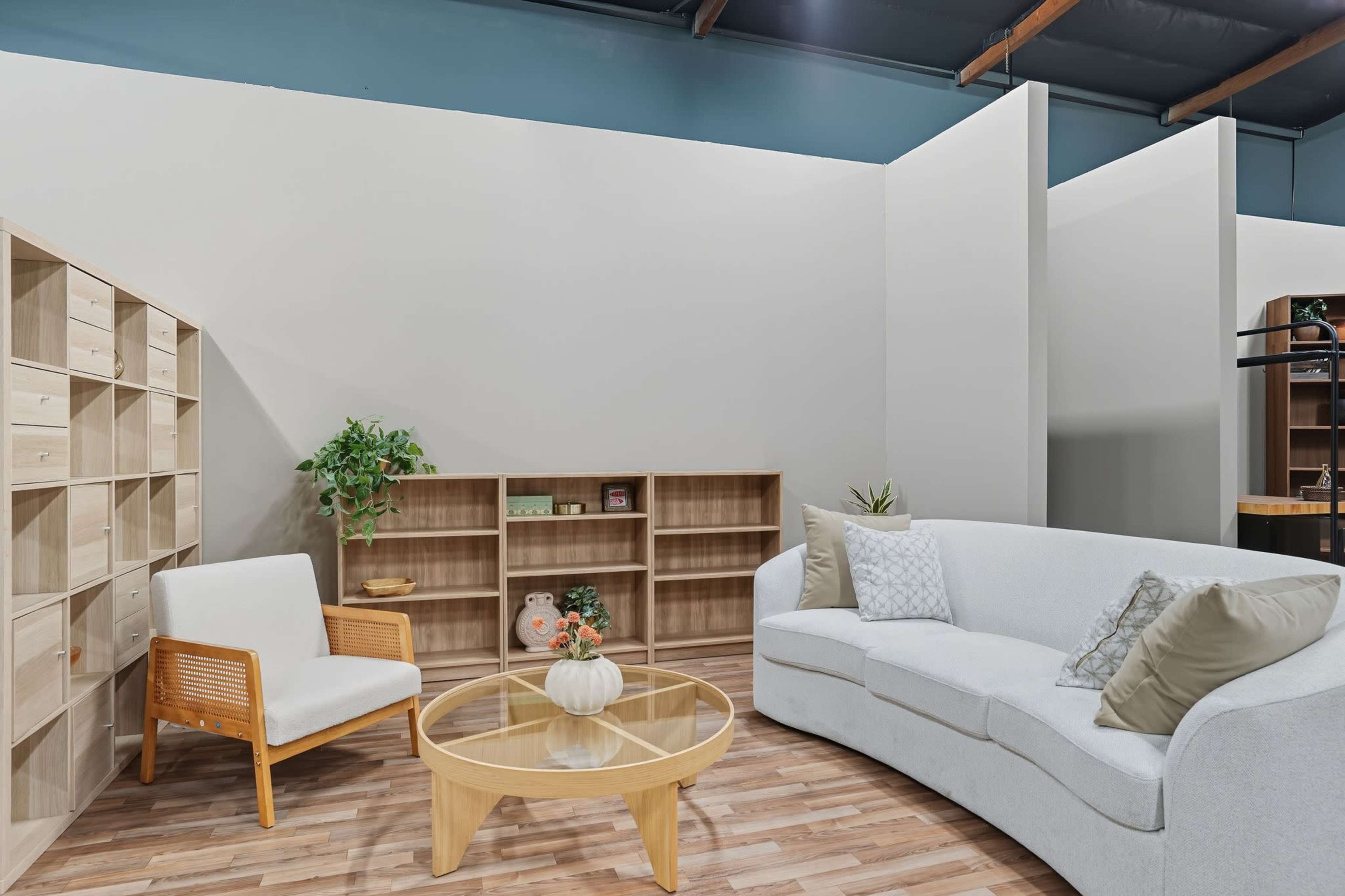 The room features a modern seating area with a curved white sofa, a glass coffee table, and a wooden shelving unit against a plain wall.