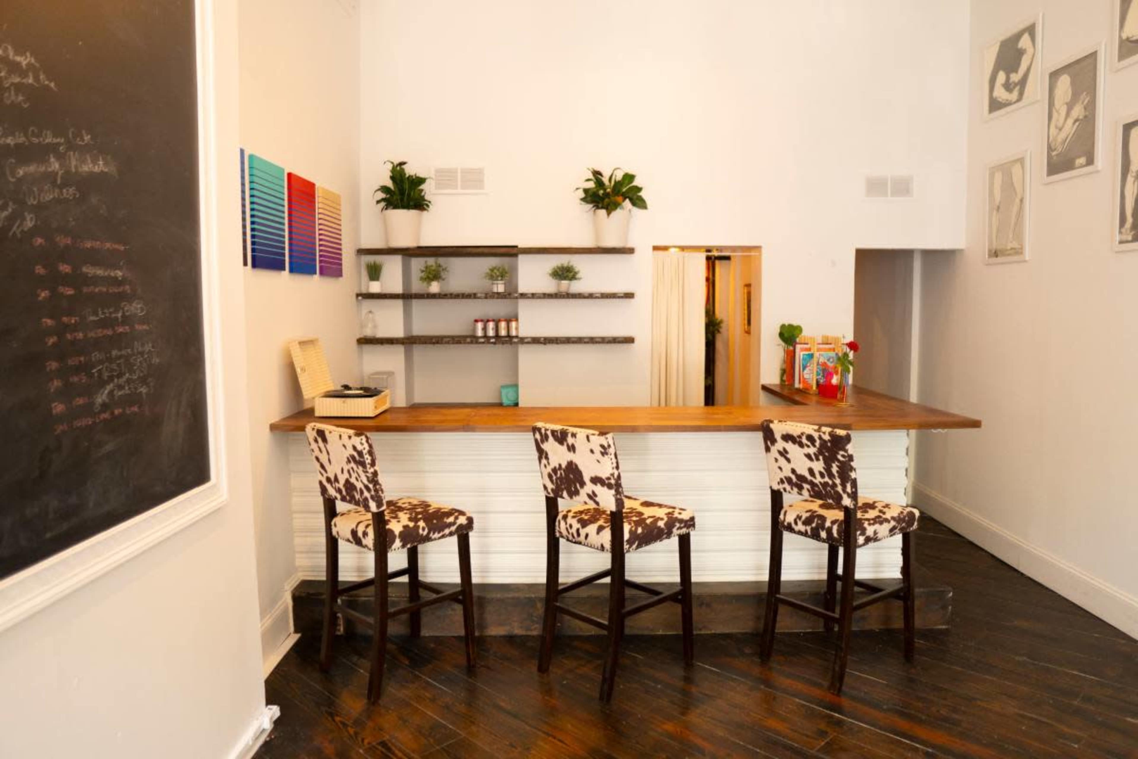 The image shows a minimalistic interior of a café or bar featuring a wooden counter, three patterned stools, a wall-mounted shelf with plants, and a chalkboard on the left.