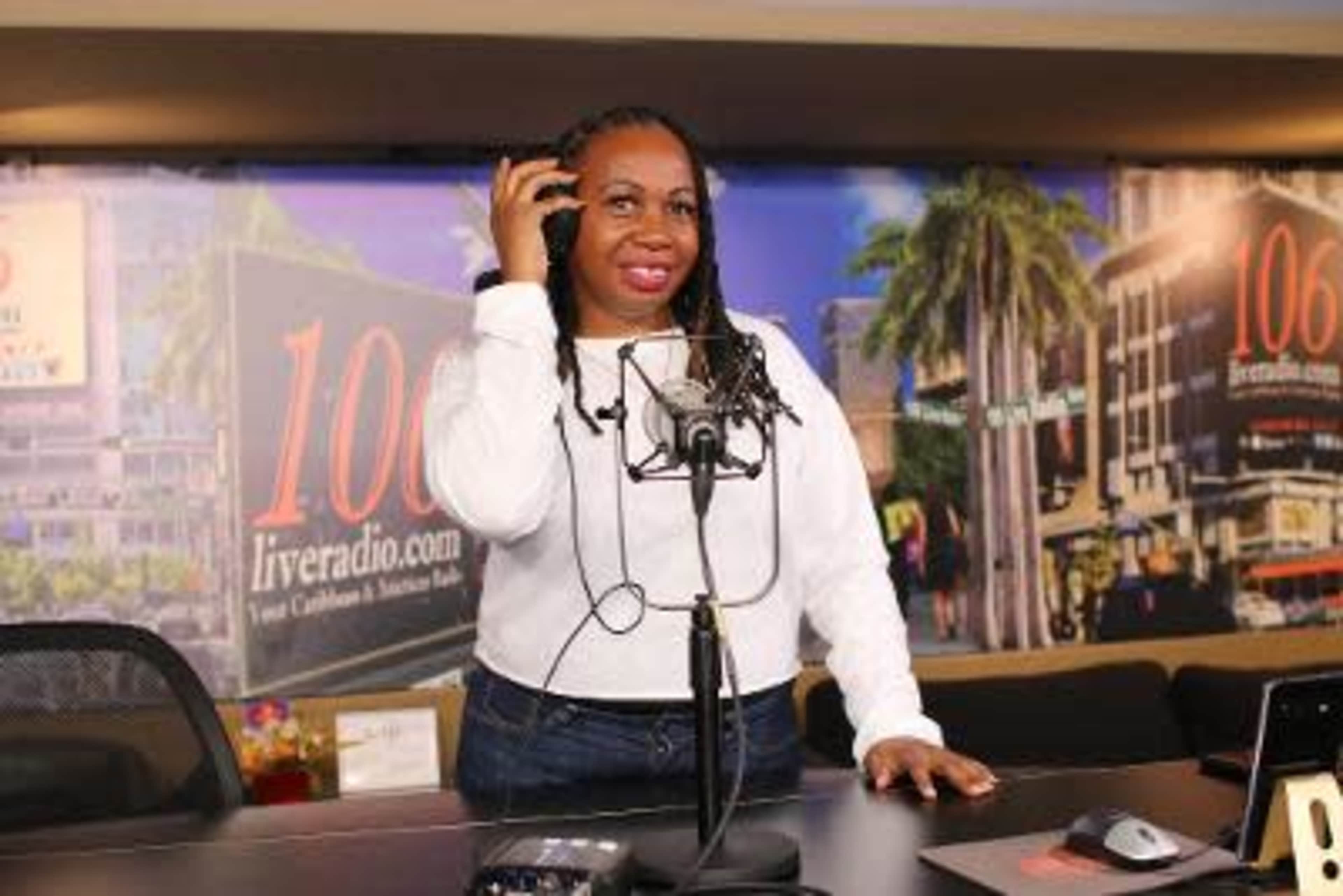 A woman stands at a radio station studio microphone, adjusting her headset with a backdrop of a promotional banner.