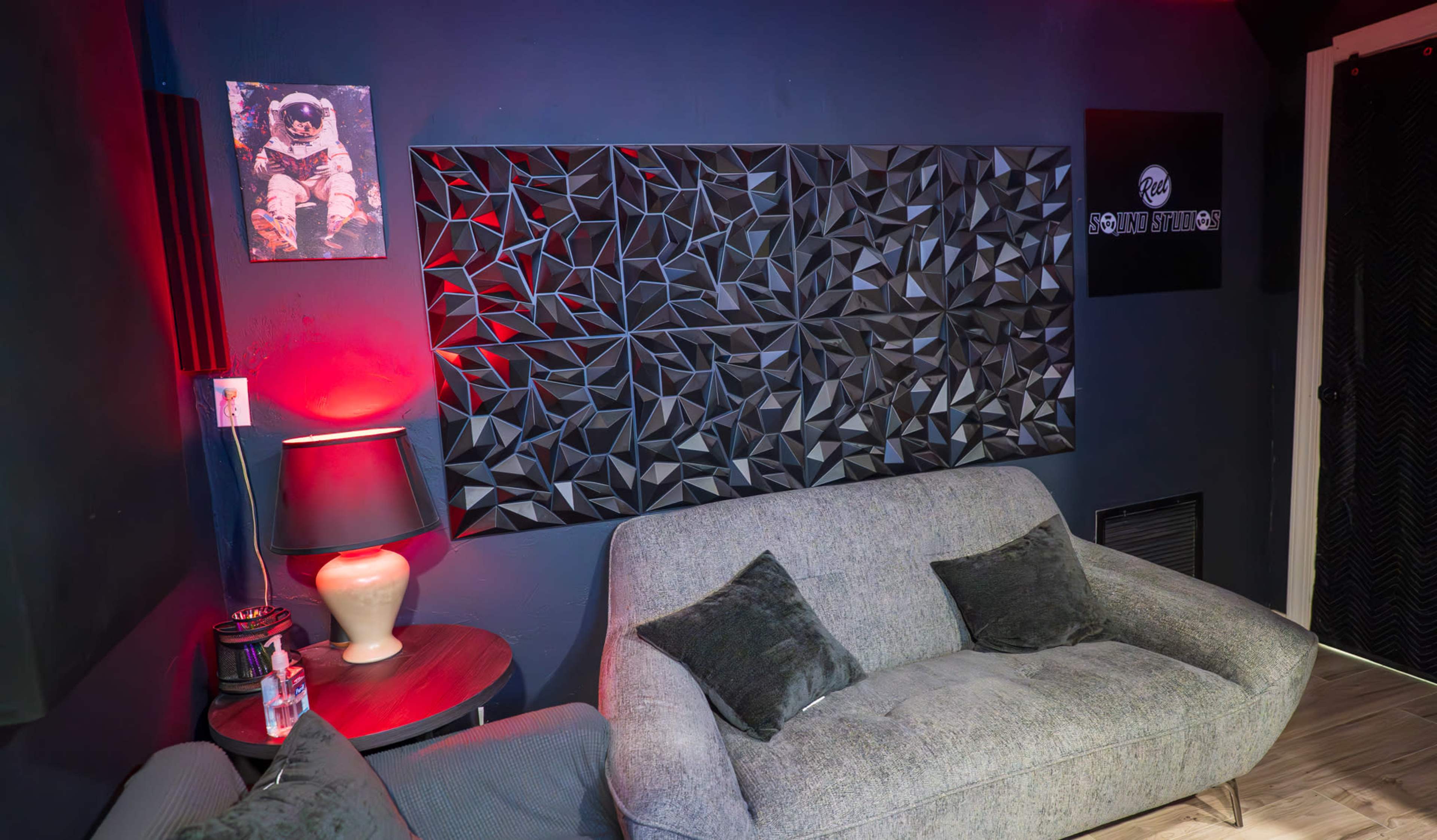The image shows a modern lounge area featuring a grey couch, a table lamp, and a textured black wall panel adorned with geometric patterns.