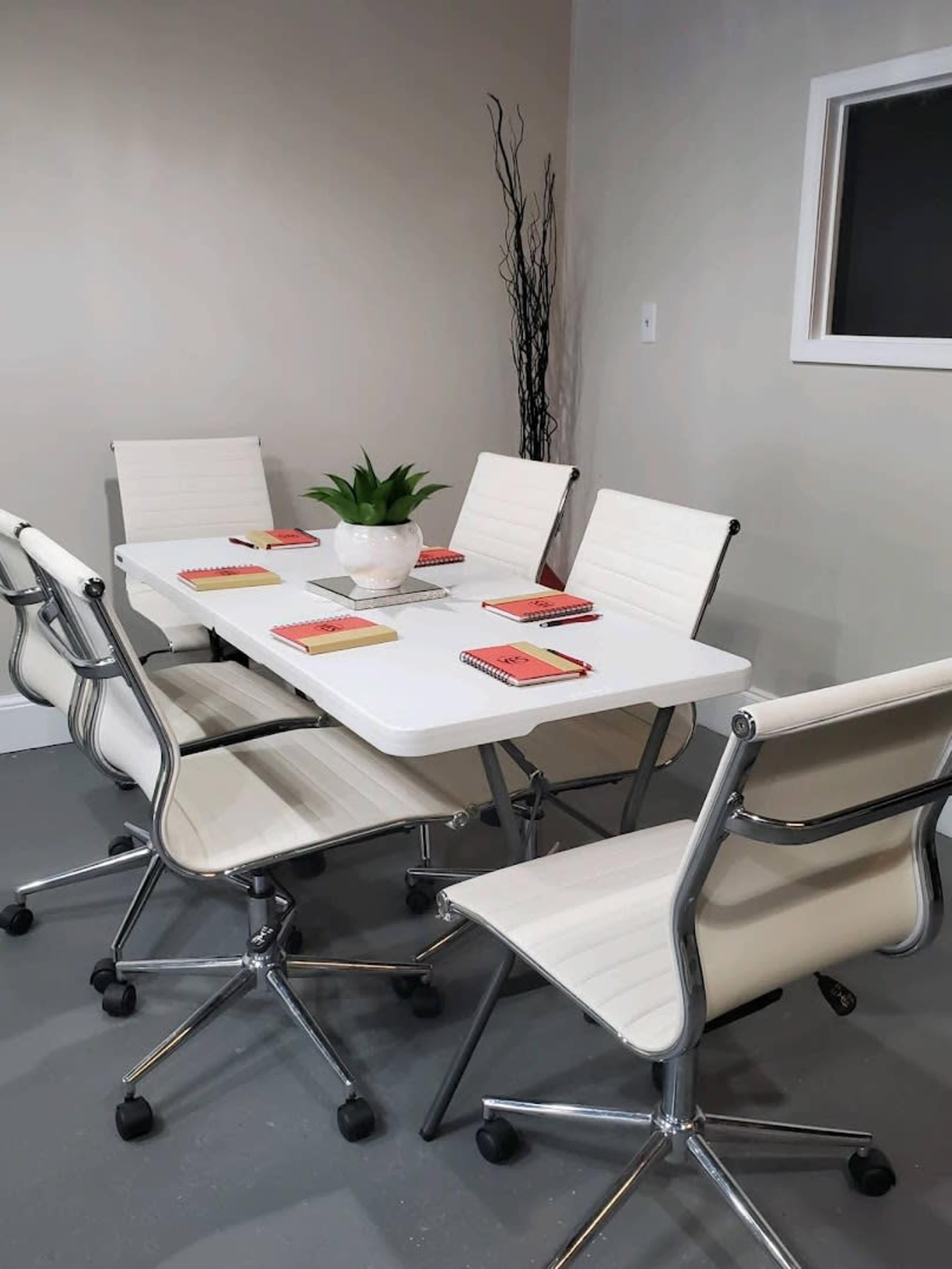 A light-colored conference room features a rectangular table surrounded by six white, modern swivel chairs, with a small plant and notepads placed at each seat.