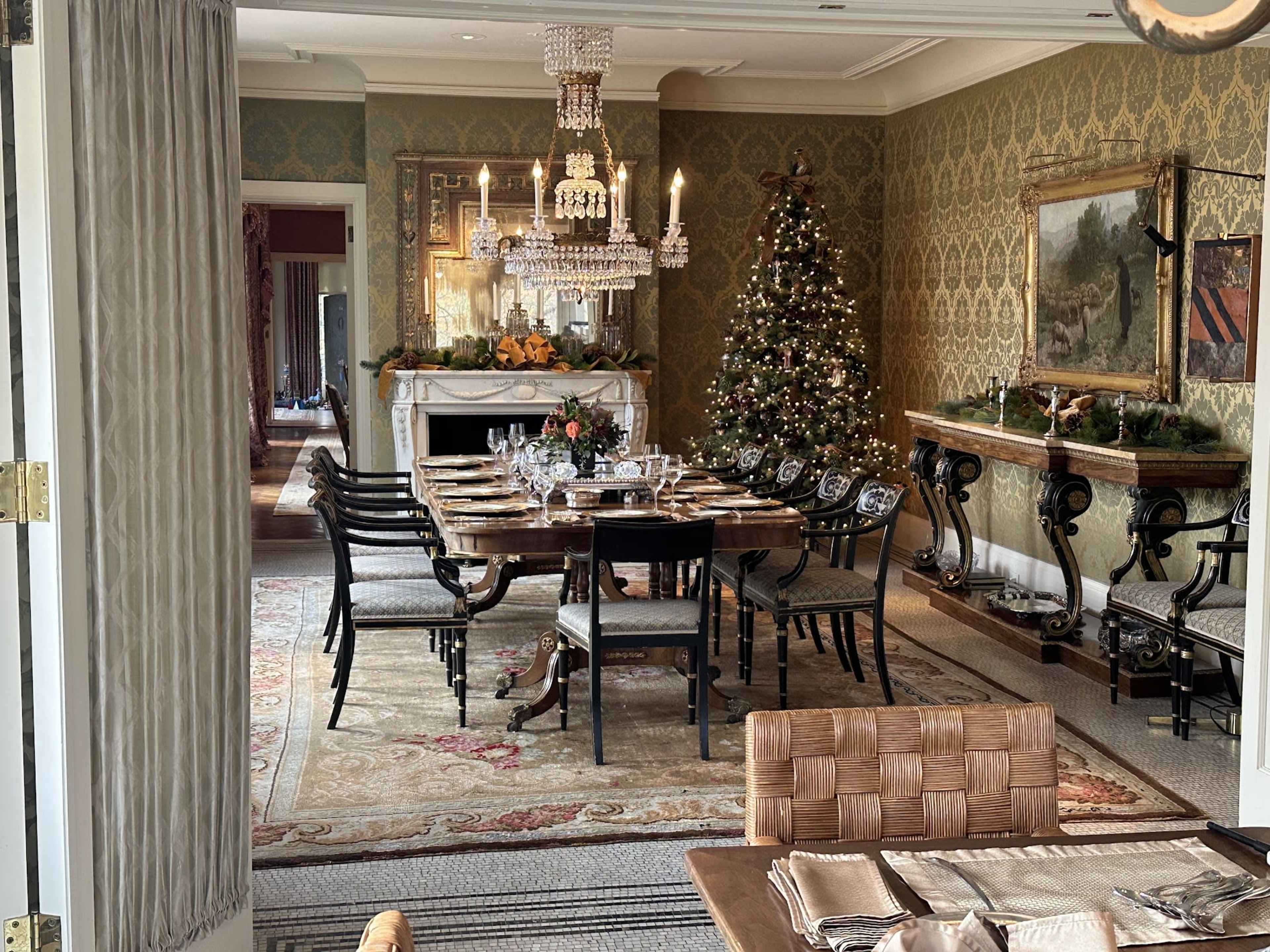 A dining room features a long wooden table set for a meal, a chandelier overhead, and a decorated Christmas tree in the corner.