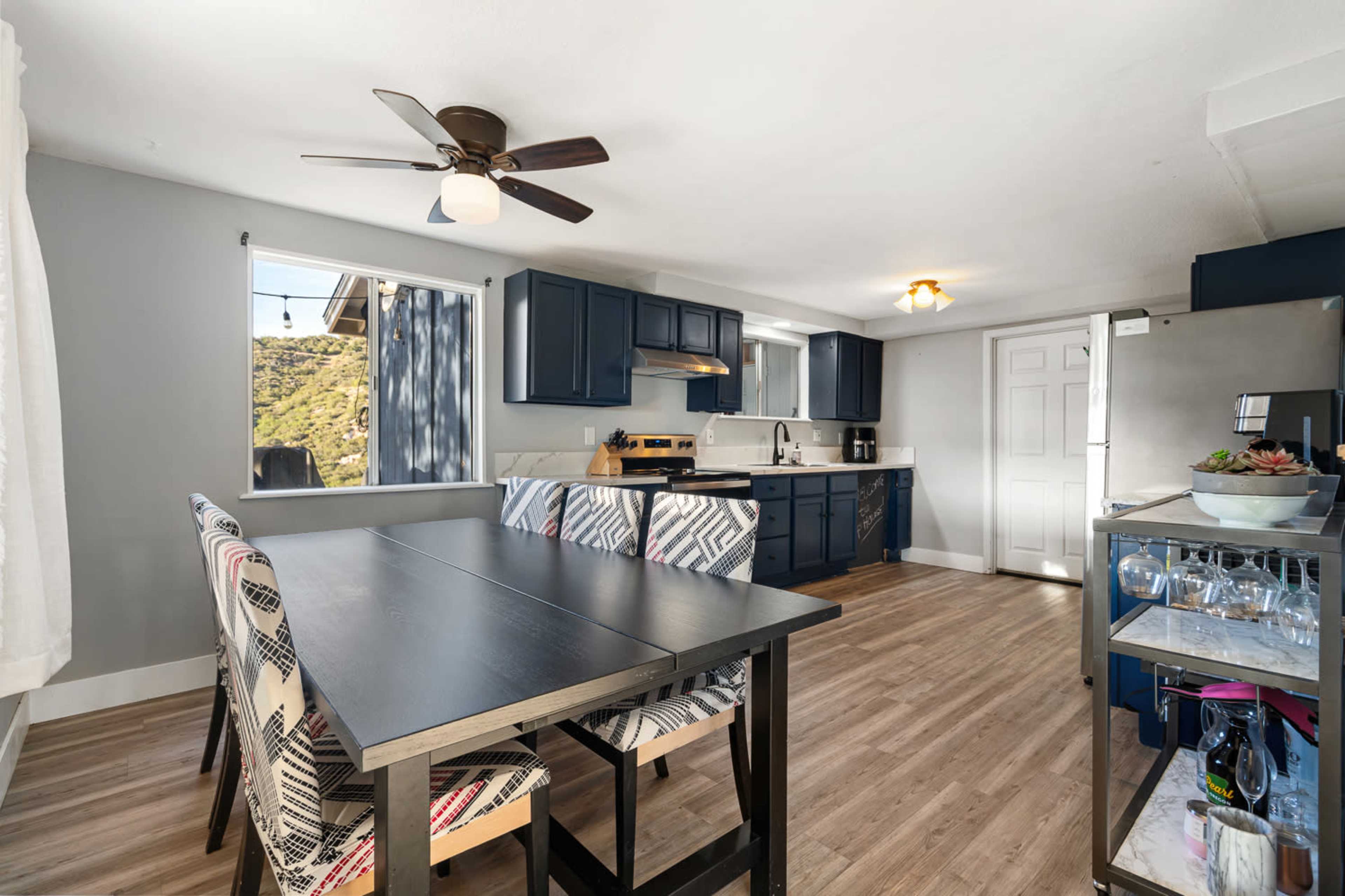 The image shows a modern kitchen and dining area featuring a table with four patterned chairs, dark blue cabinets, and vinyl flooring.