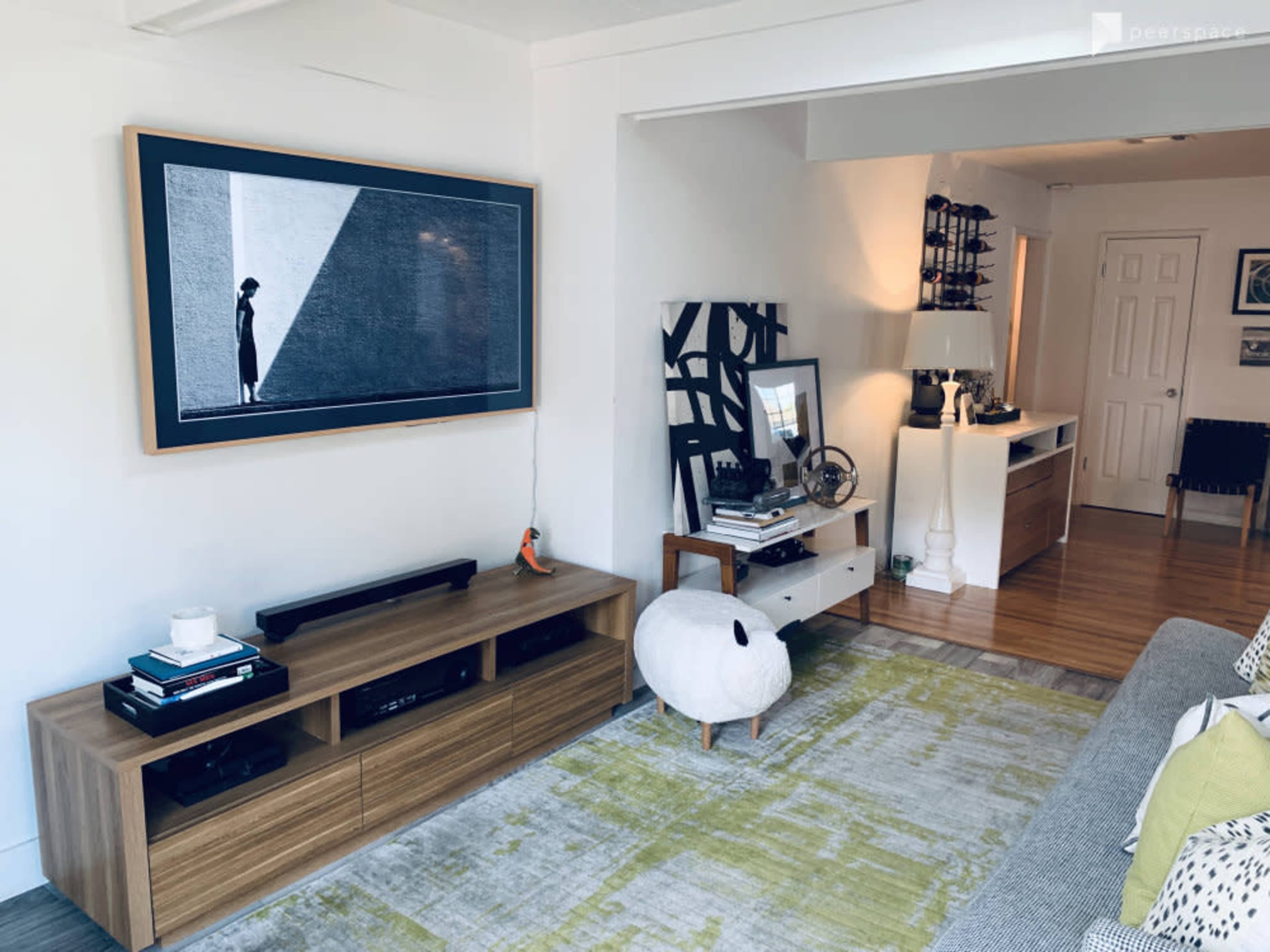 A living room features a large television mounted on the wall, a wooden media console, a patterned green and white rug, and various decorative items on the furniture.
