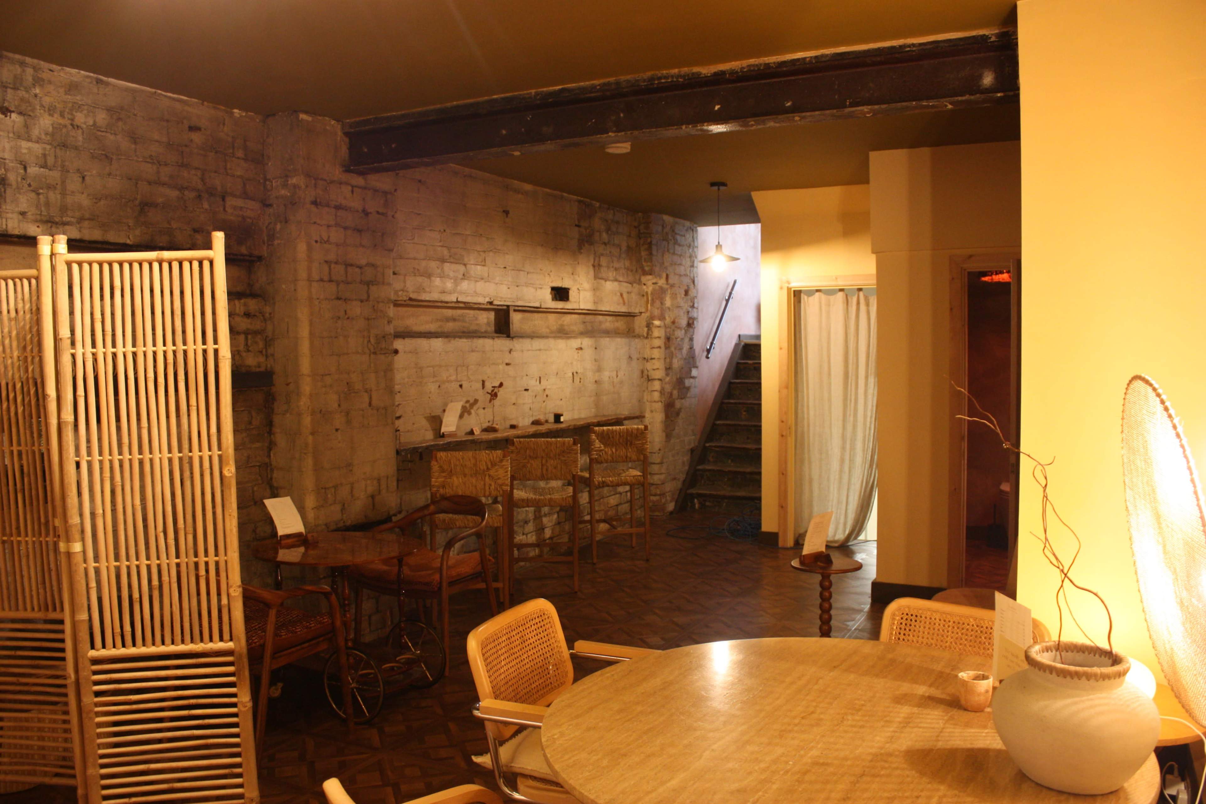 A spacious, minimalist interior with exposed brick walls, a wooden beam ceiling, and a staircase leading to another area, illuminated by soft lighting.