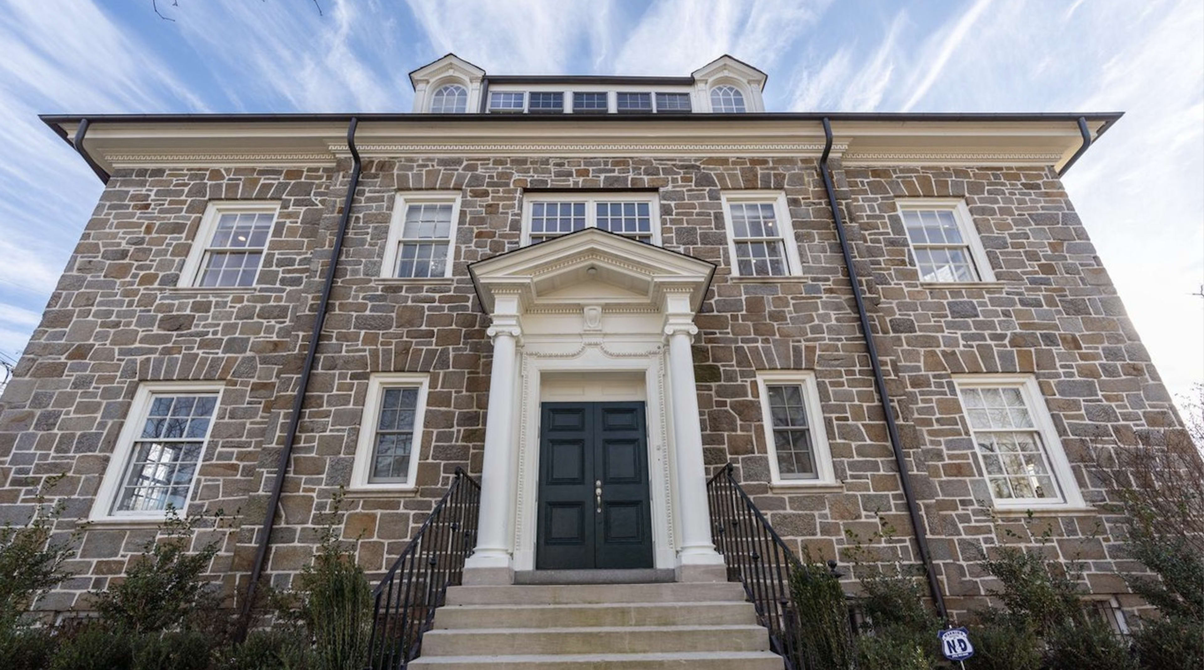 A large stone building features a central entrance with a white portico and stairs leading up to a double green door.