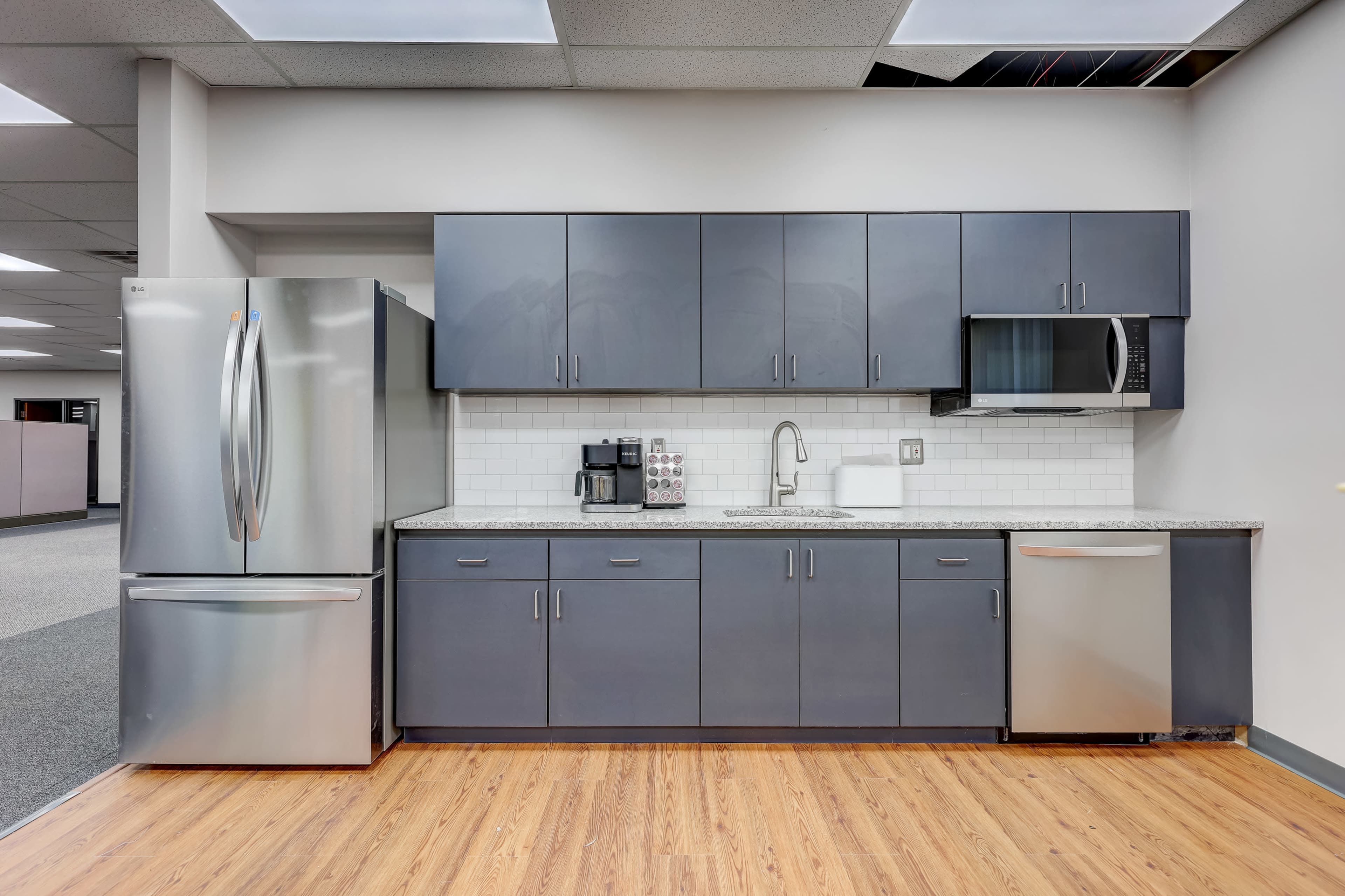 A kitchen with dark gray cabinets, a stainless steel refrigerator, a microwave, and a sink, all set against a light tile backsplash and wooden flooring.