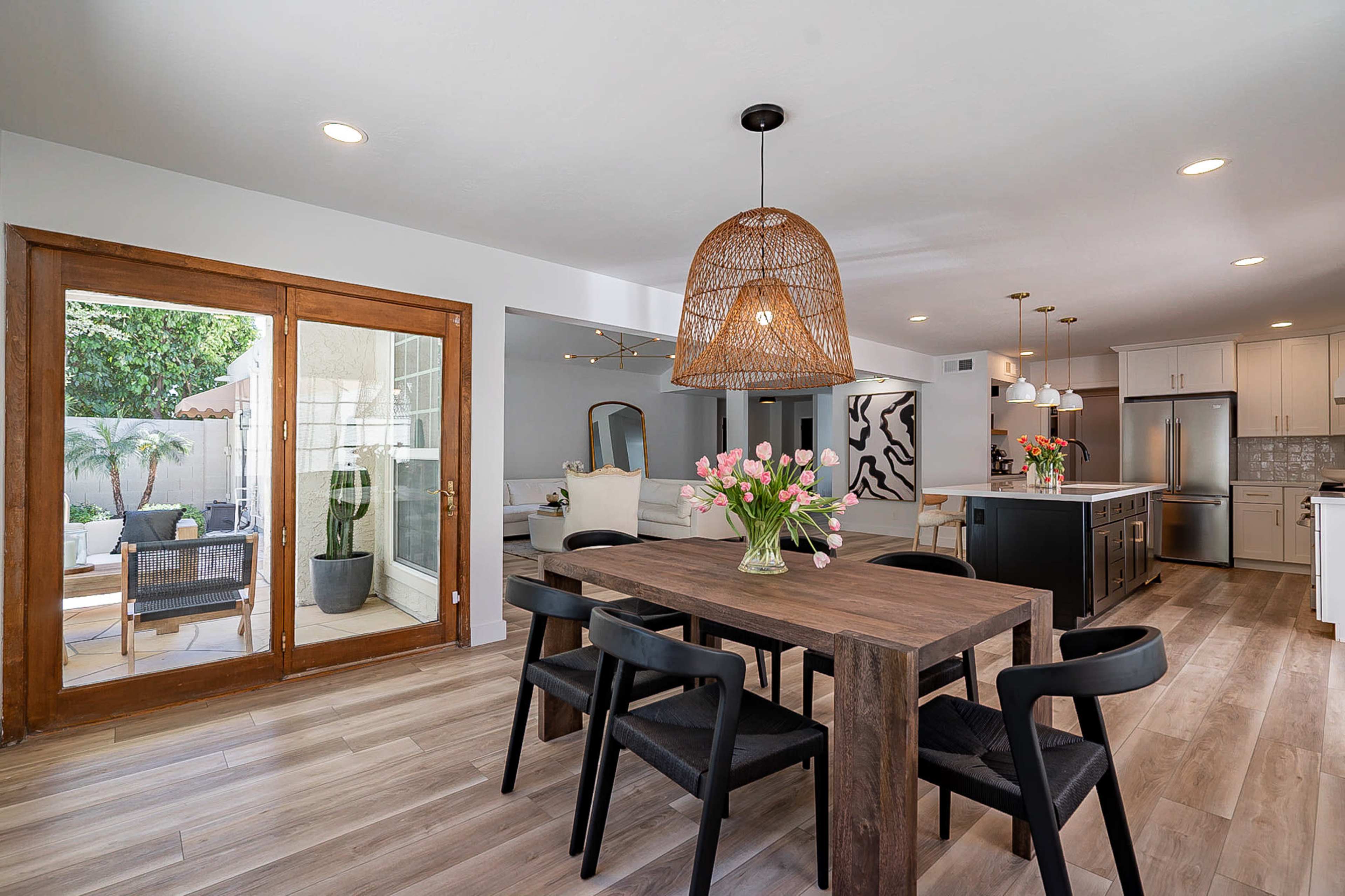 A dining area features a wooden table with black chairs, surrounded by natural light from large glass doors leading to an outdoor space.
