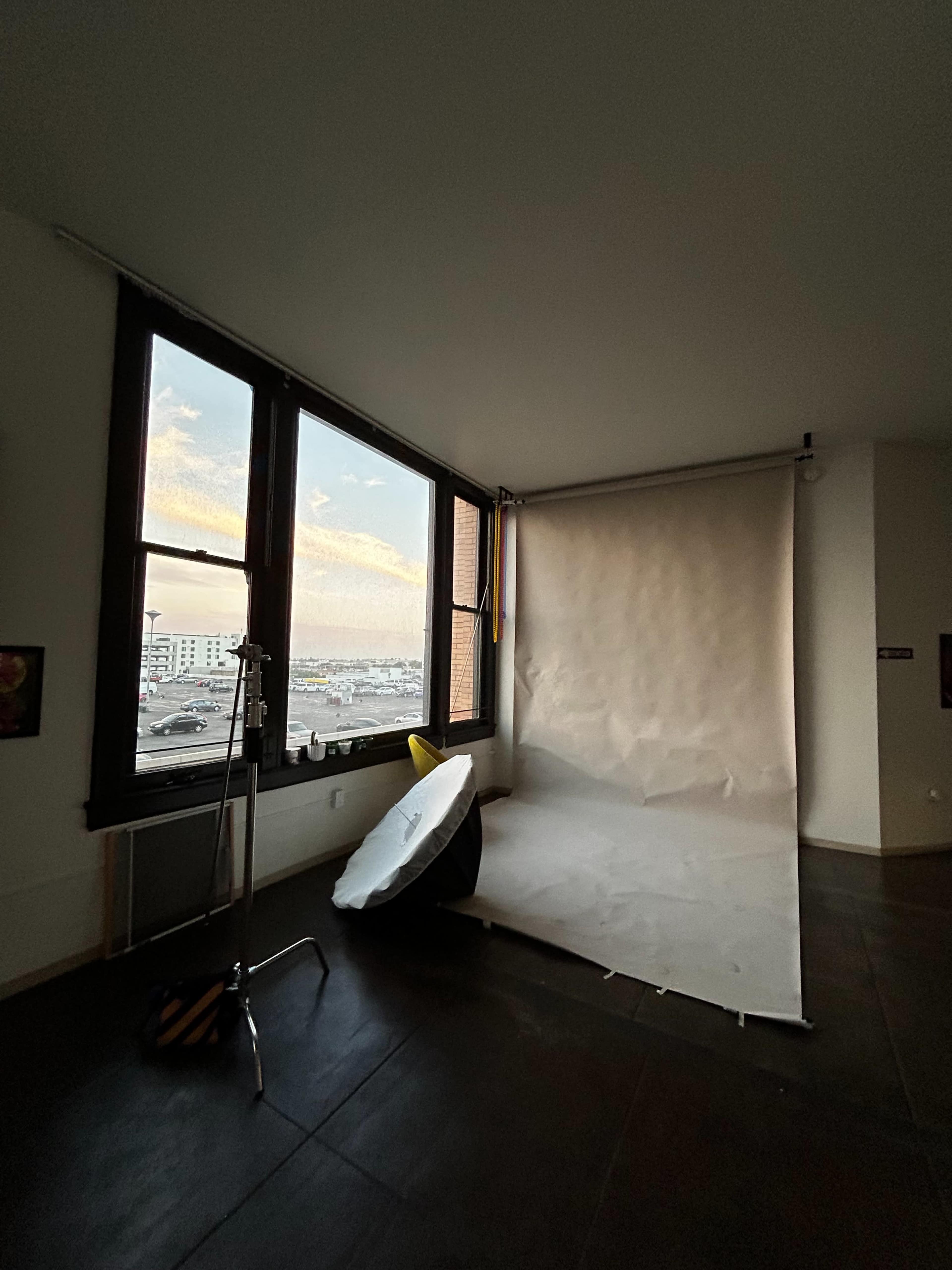 The image shows a photography studio with a white backdrop, a light reflector, and large windows revealing an urban skyline at dusk.