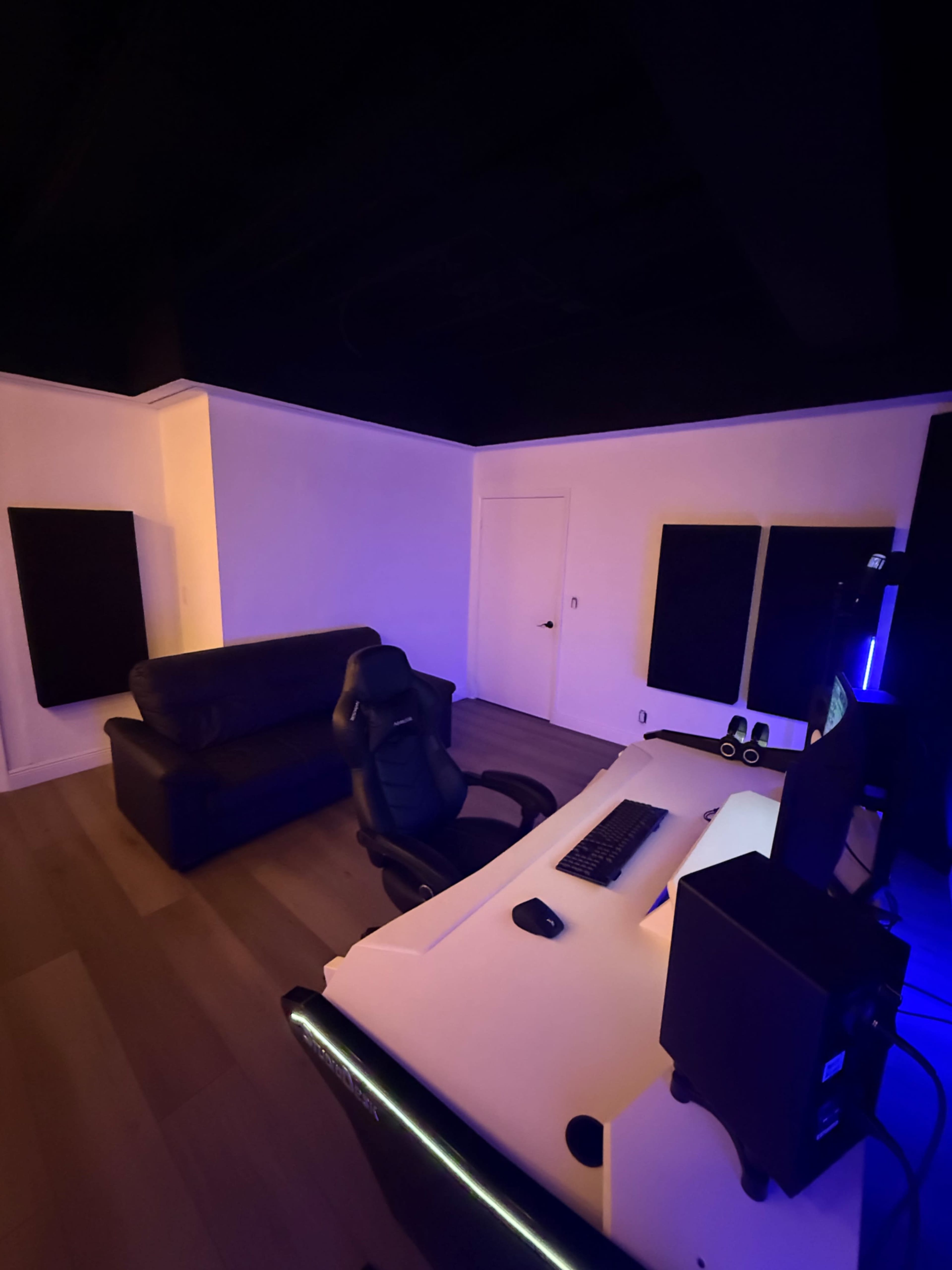 The image shows a modern room with a black sofa, a sleek desk with dual monitors, and soundproof panels on the walls, illuminated by colored lighting.