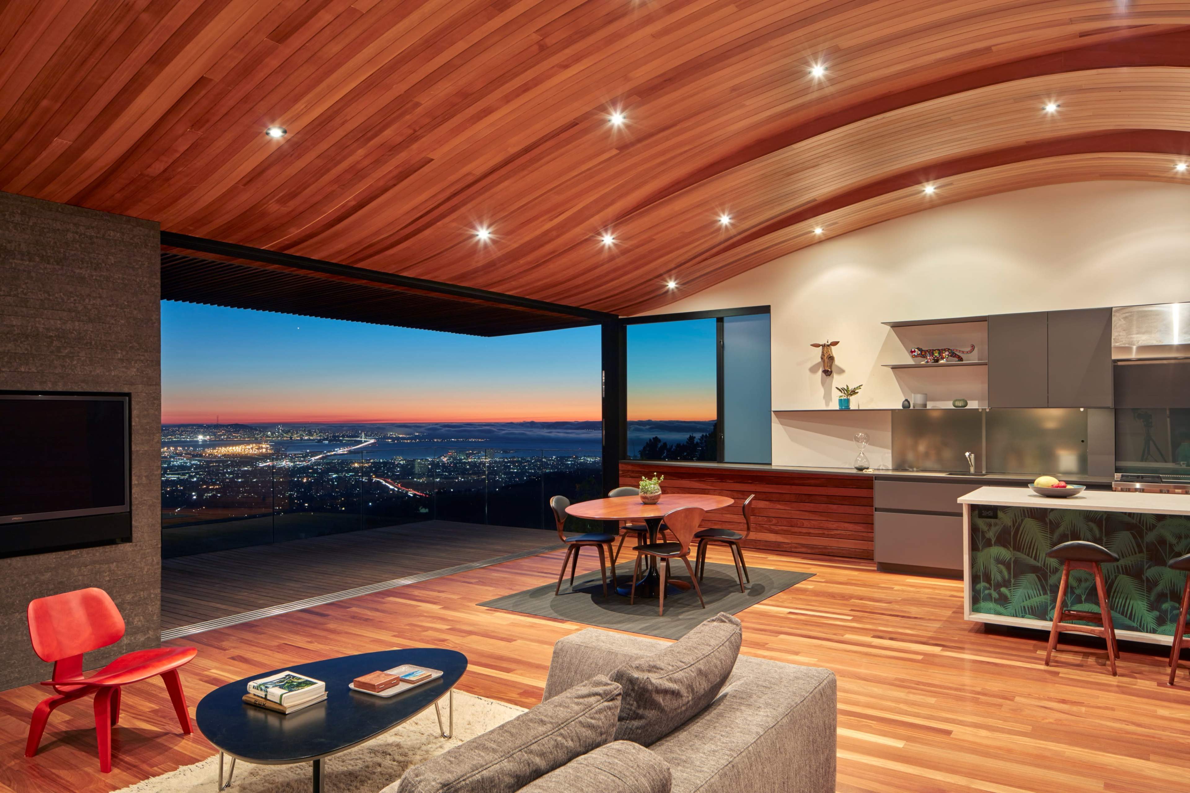 A modern living space features a spacious open layout with wooden ceilings, a kitchen, a dining area, and large windows showcasing a city skyline at sunset.