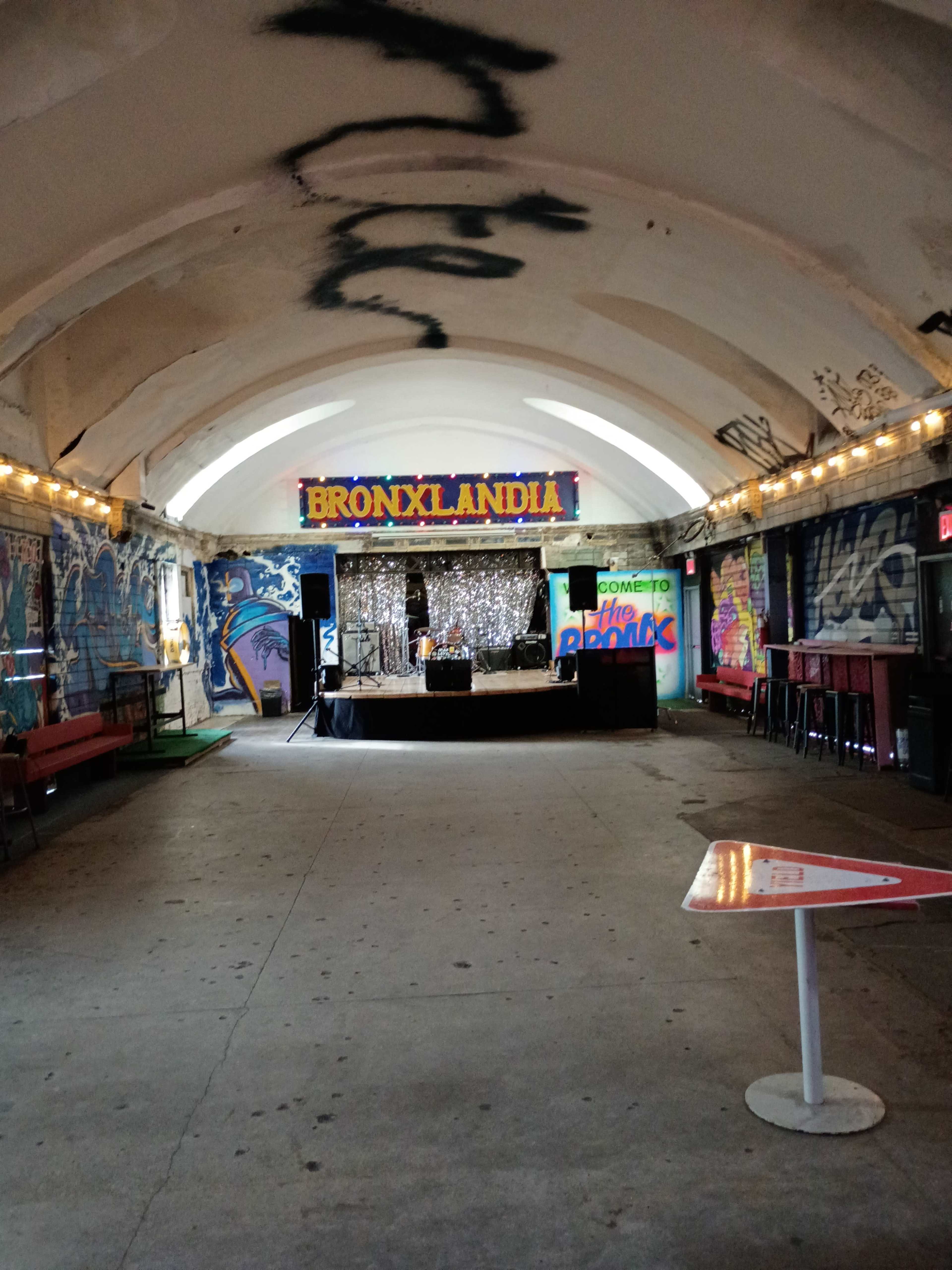 The image shows an indoor event space with colorful graffiti, a stage with decorations, and seating areas along the walls.