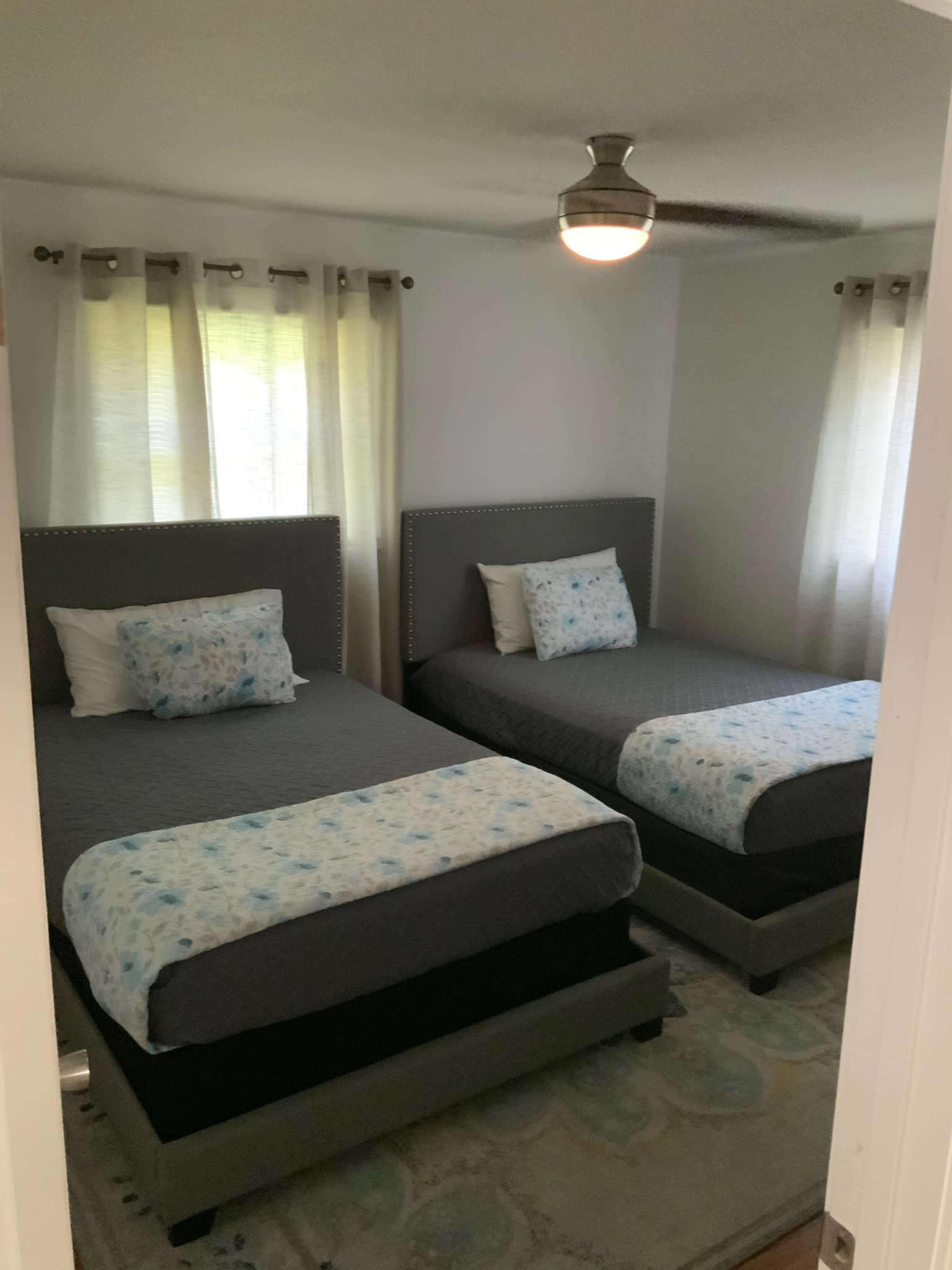 A room with two twin beds, each covered with gray bedding and topped with blue and white patterned throw pillows, next to a window with light curtains.