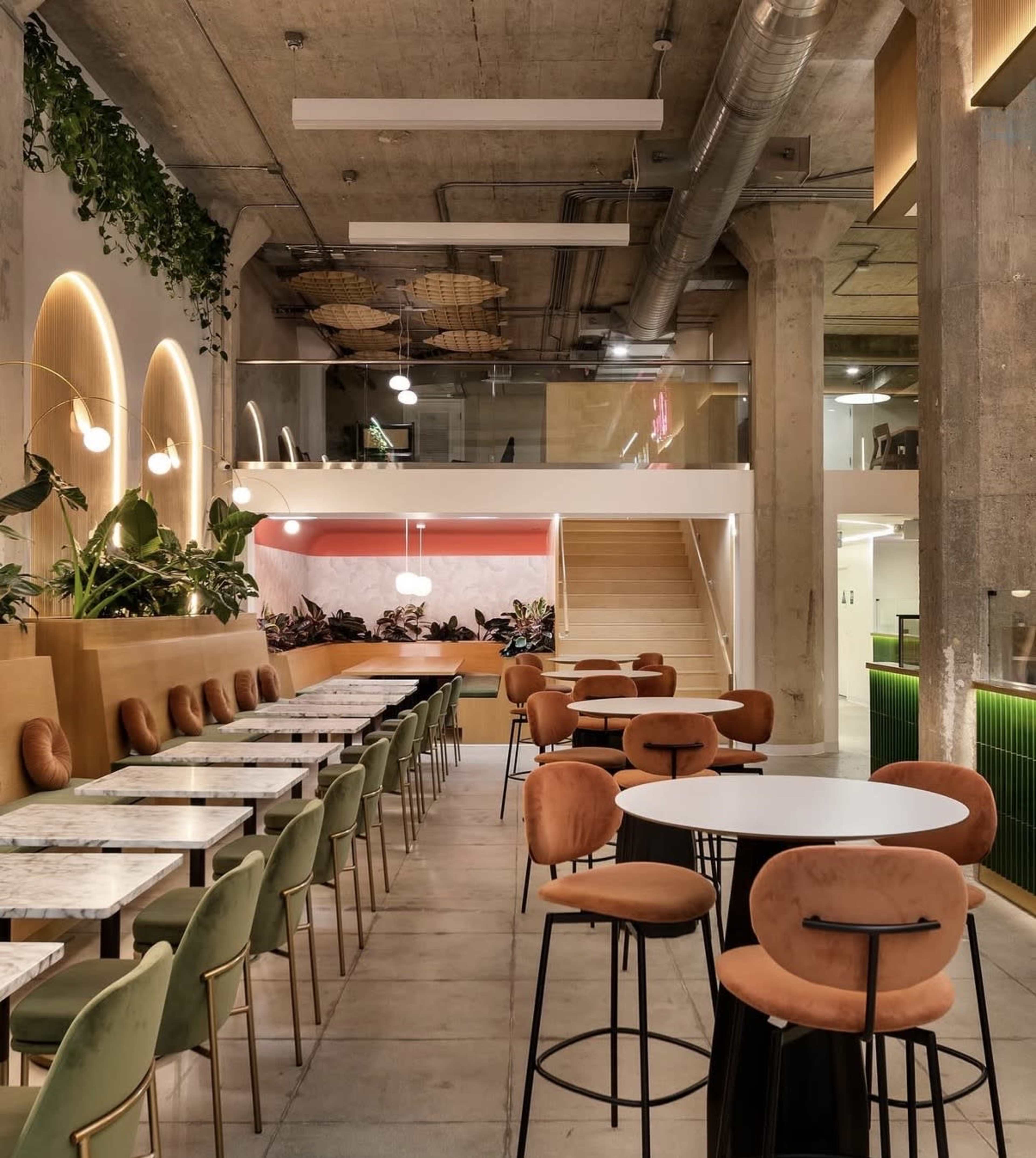The image shows a modern café interior with tables, colorful seating, and greenery, featuring a stylish staircase and decorative lighting.