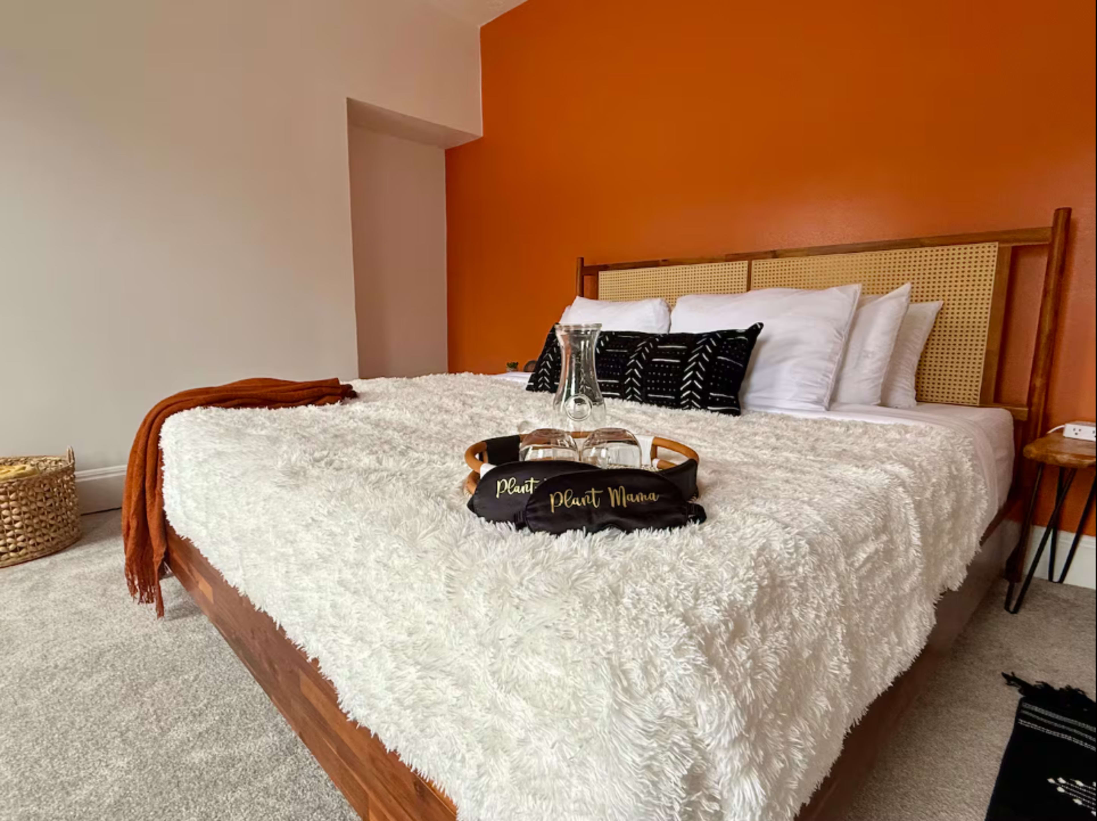 A bed with a white fuzzy blanket and black pillows is set against an orange wall in a well-lit bedroom.
