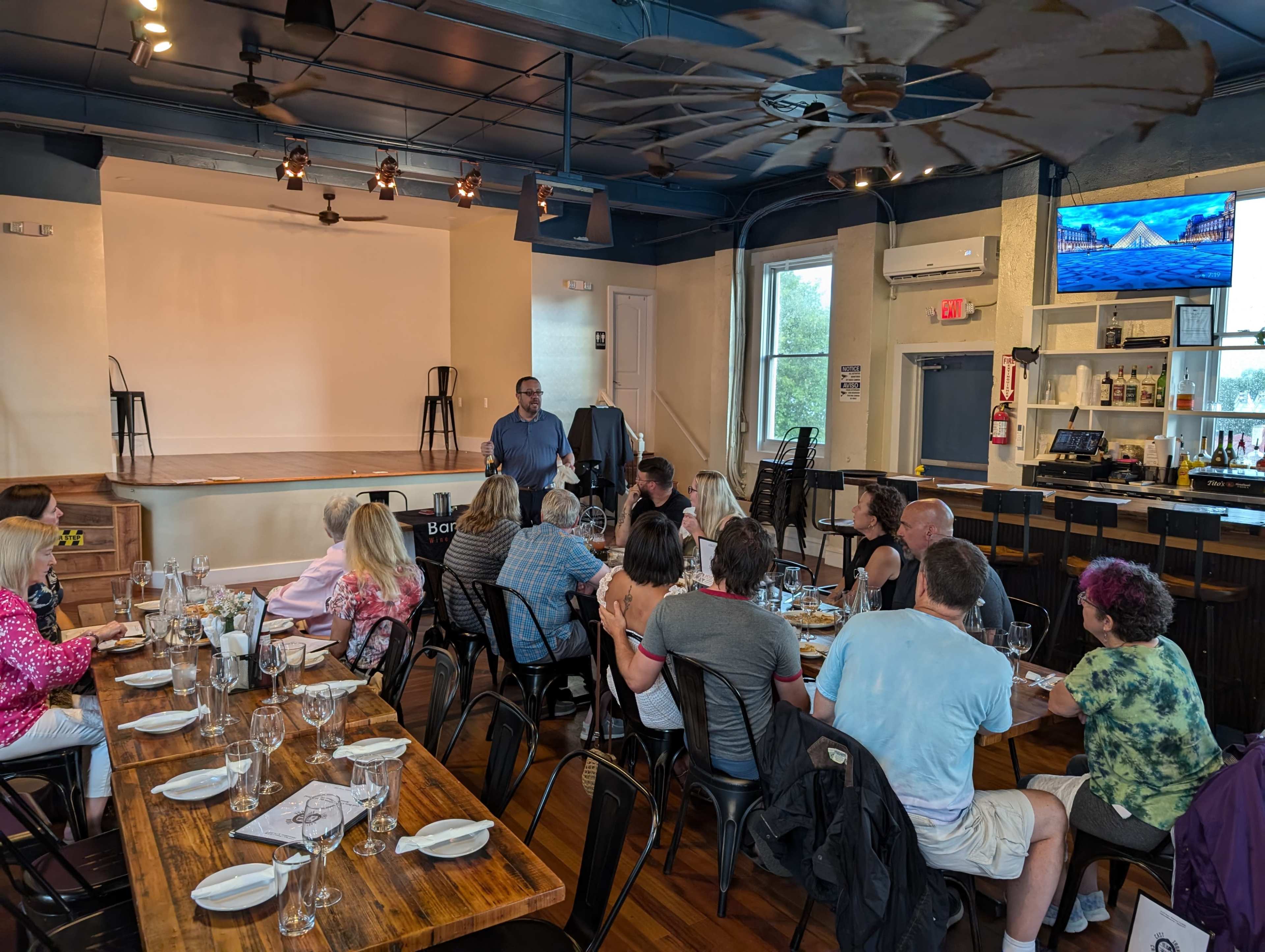 A man speaks to an audience seated at tables in a restaurant with a small stage and a bar area in the background.