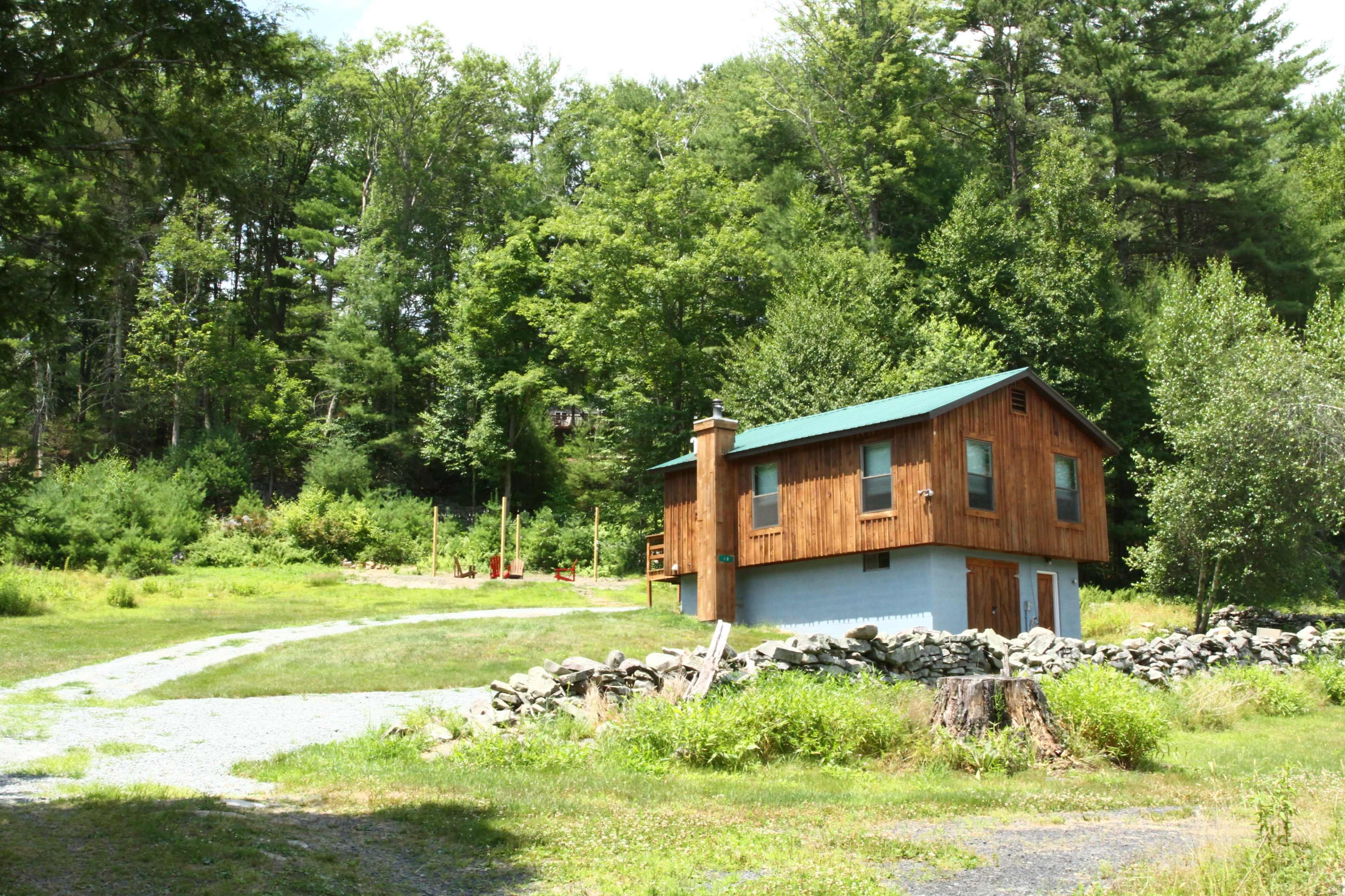 A wooden two-story cabin with a green roof is situated near a gravel path and surrounded by trees and shrubs.