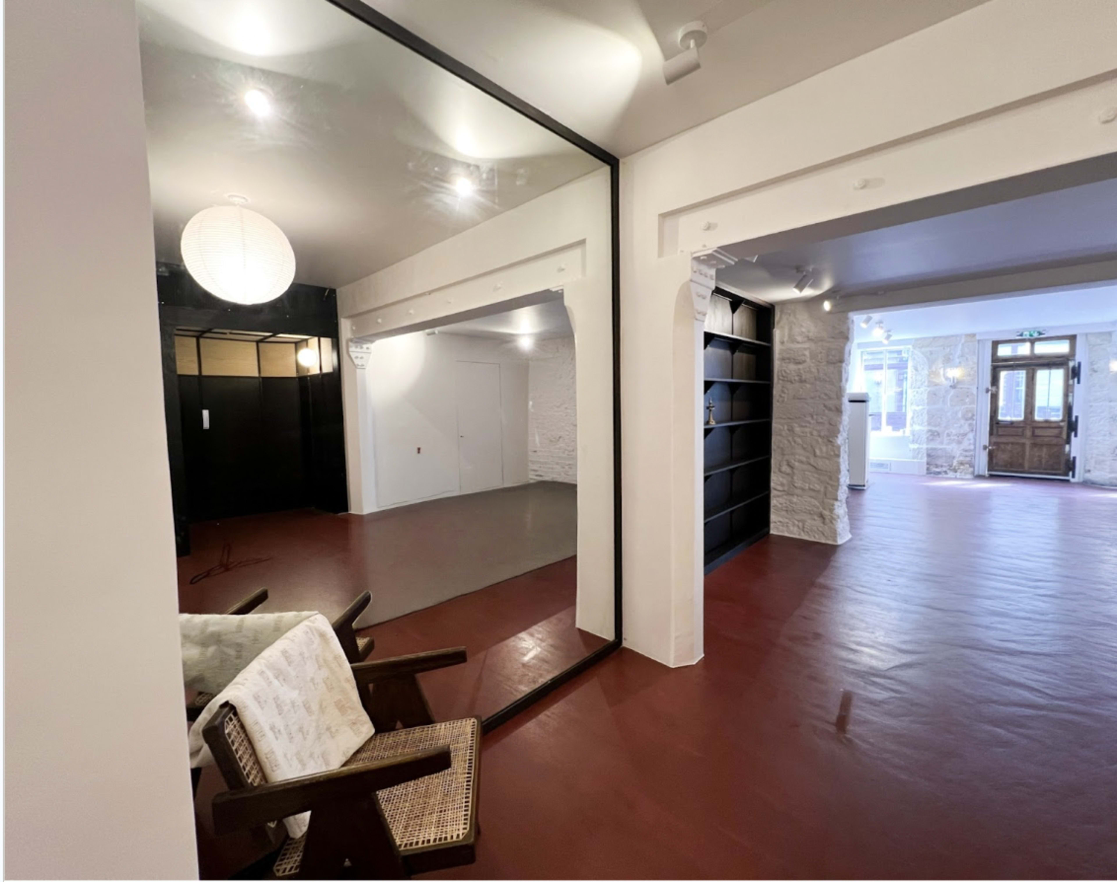 The image shows an interior space featuring a large mirror, a wicker chair, and a light-colored wall, leading to another room with stone walls and a wooden entrance.
